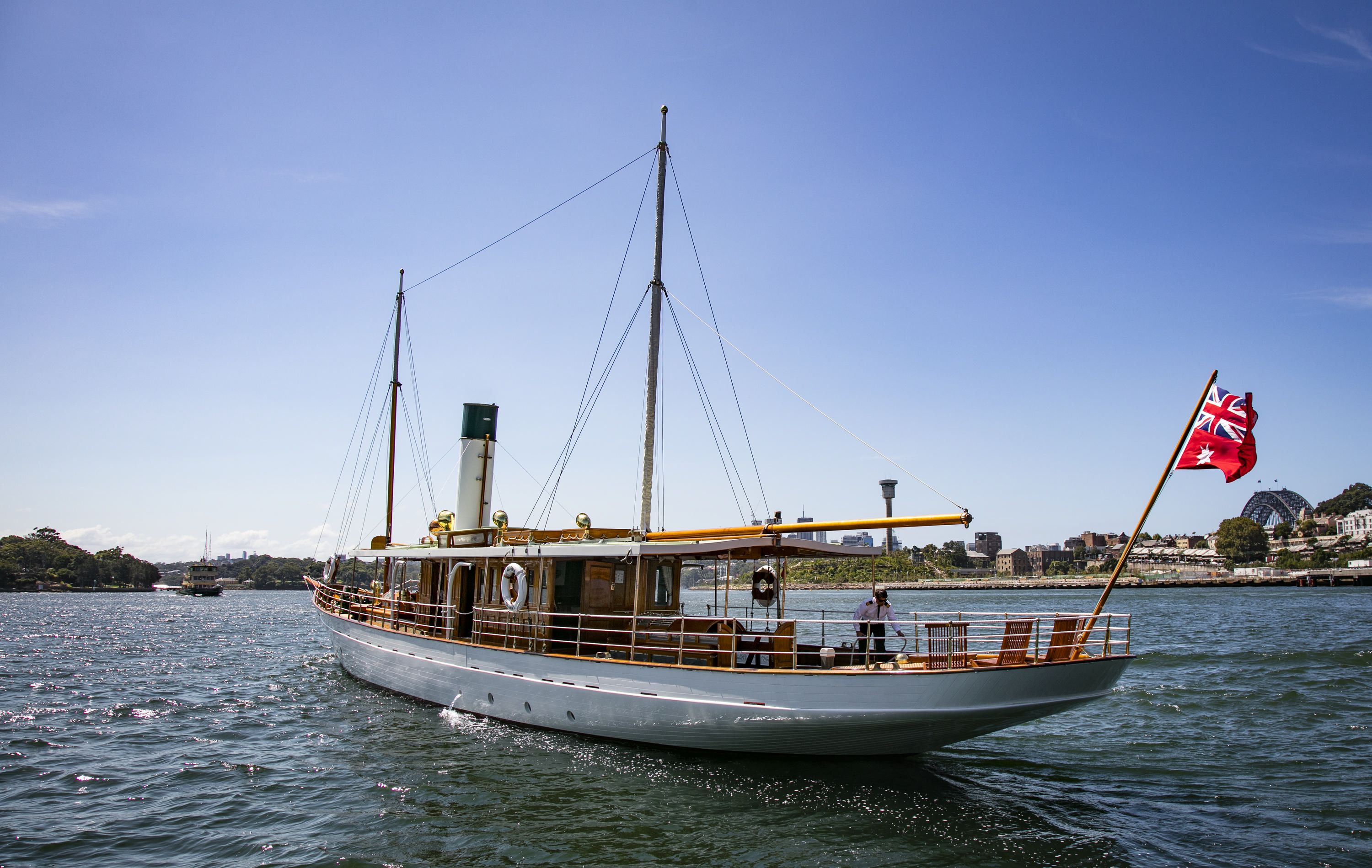 Photo of Edwardian steam yacht ENA out on the water, Darling Harbour - Sydney. It is a sunny day with blue sky and water, a flay is flying from the rear of the white and timber boat and the harbour bridge can be seen in the background. 