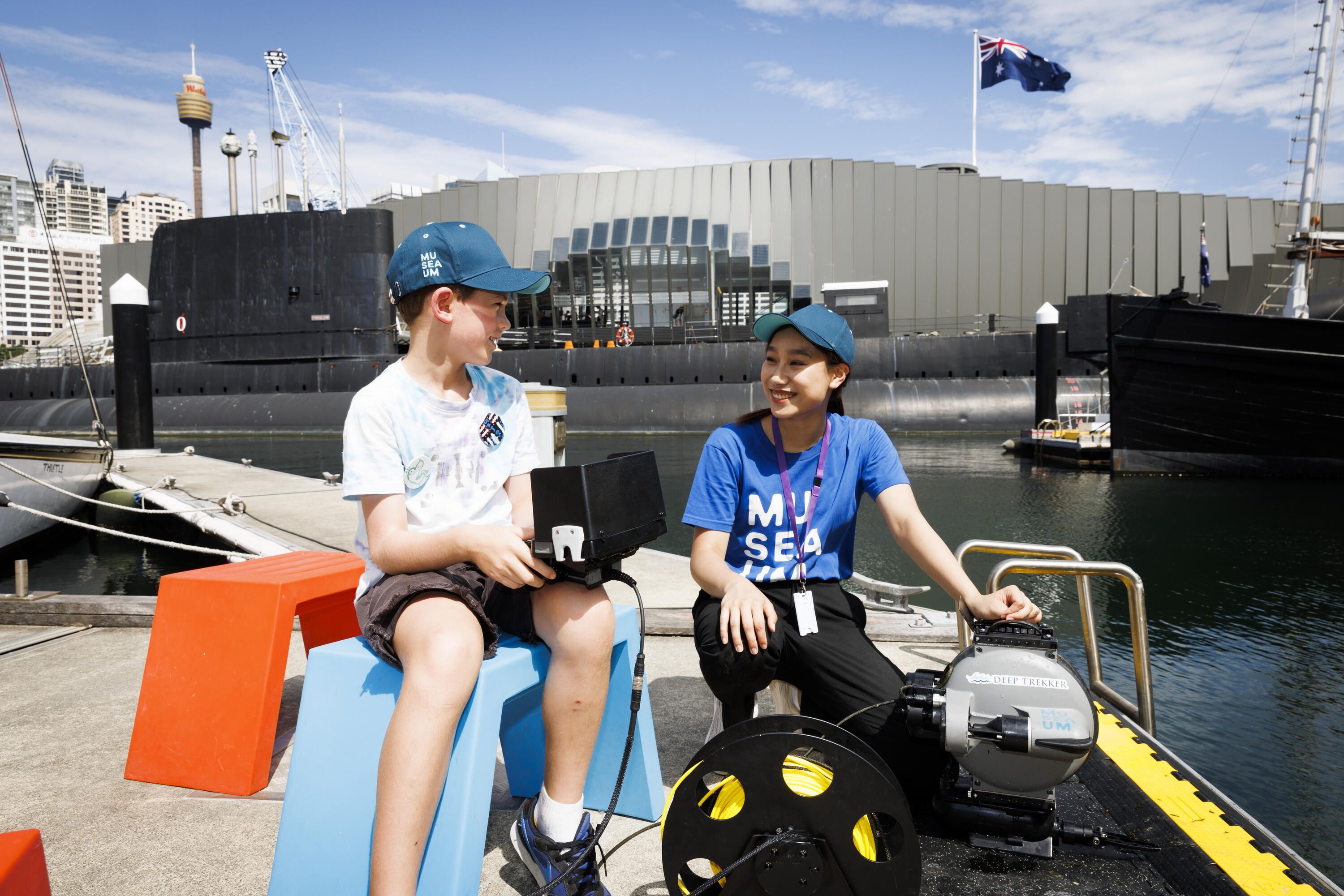Photo of a boy wearing a blue cap and a woman wearing a blue shirt with the museum logo. they are sitting outside on a wharf, the museum building in the background. 