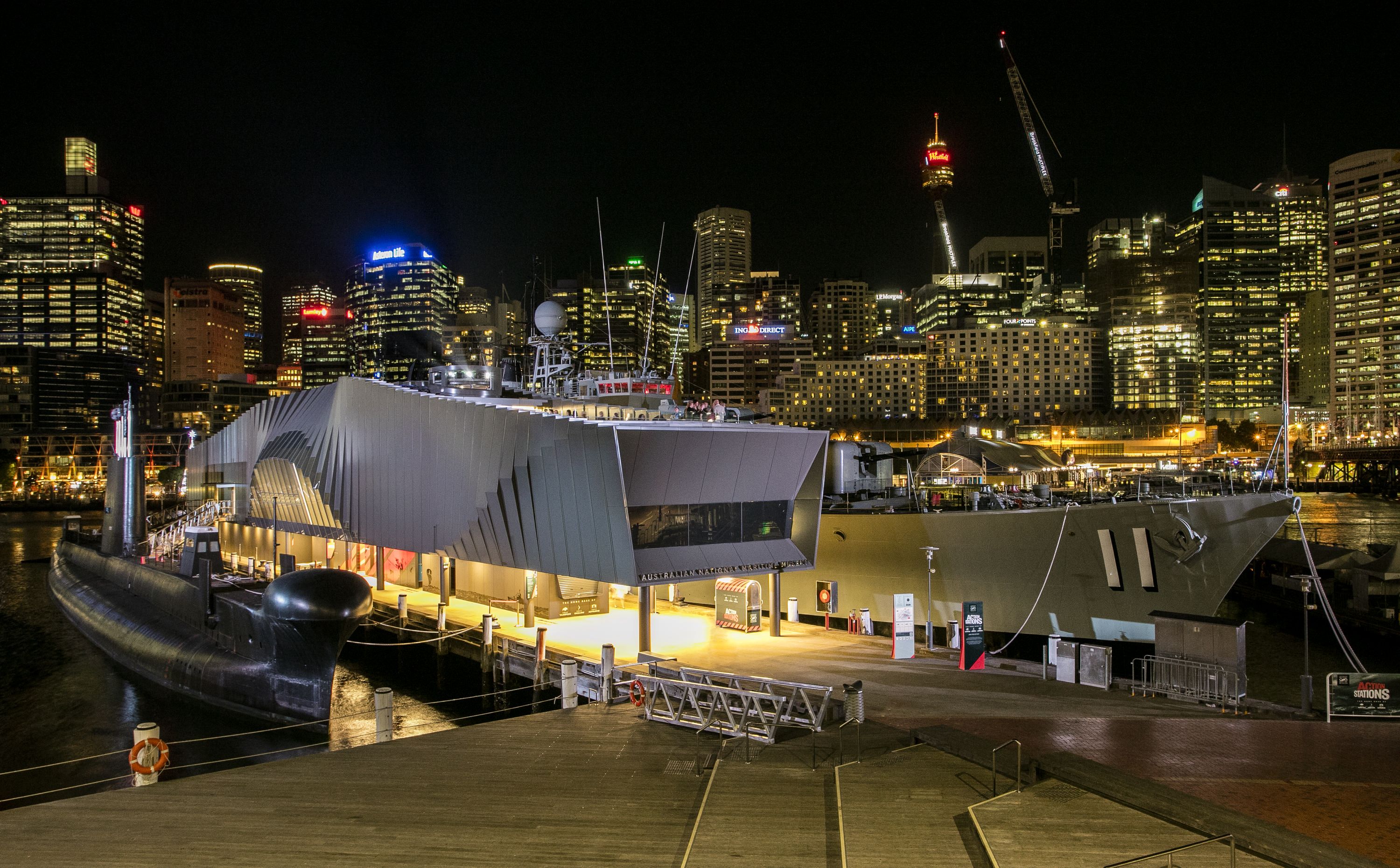 Exterior views of the Waterfront Pavilion taken at night, with the submarine HMAS ONSLOW moored at left and HMAS VAMPIRE on the right. Illuminated city buildings appear in the background.