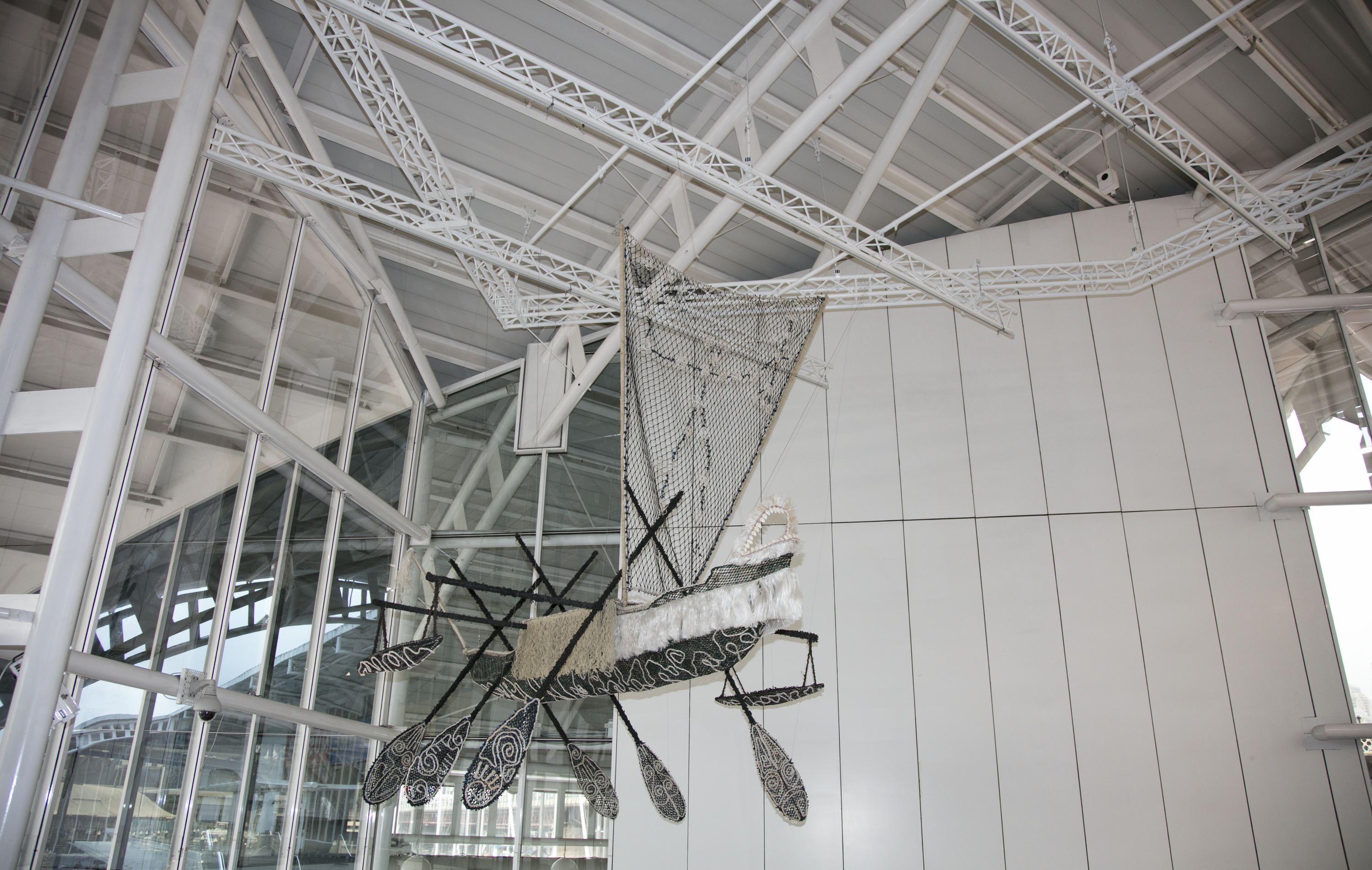 Sculpture in the shape of a boat with a sail and paddles, made of fishing nets and rope hanging from the roof. 