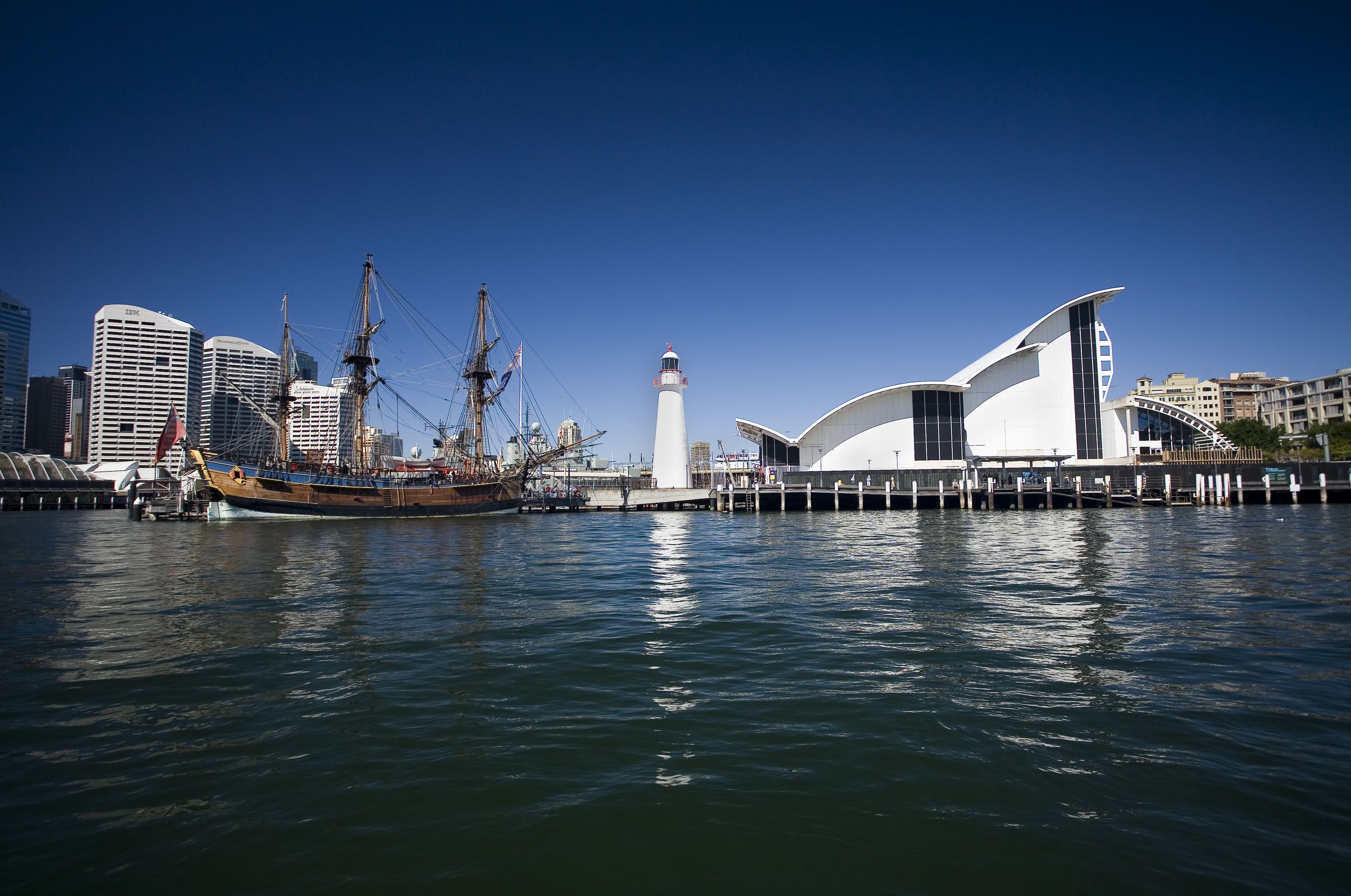 A wide landscape view of the museum building in daytime with clear blue skies. The building is shown with it's arches in profile with Cape Bowling Green lighthouse and the ENDEAVOUR replica tall ship on left.
