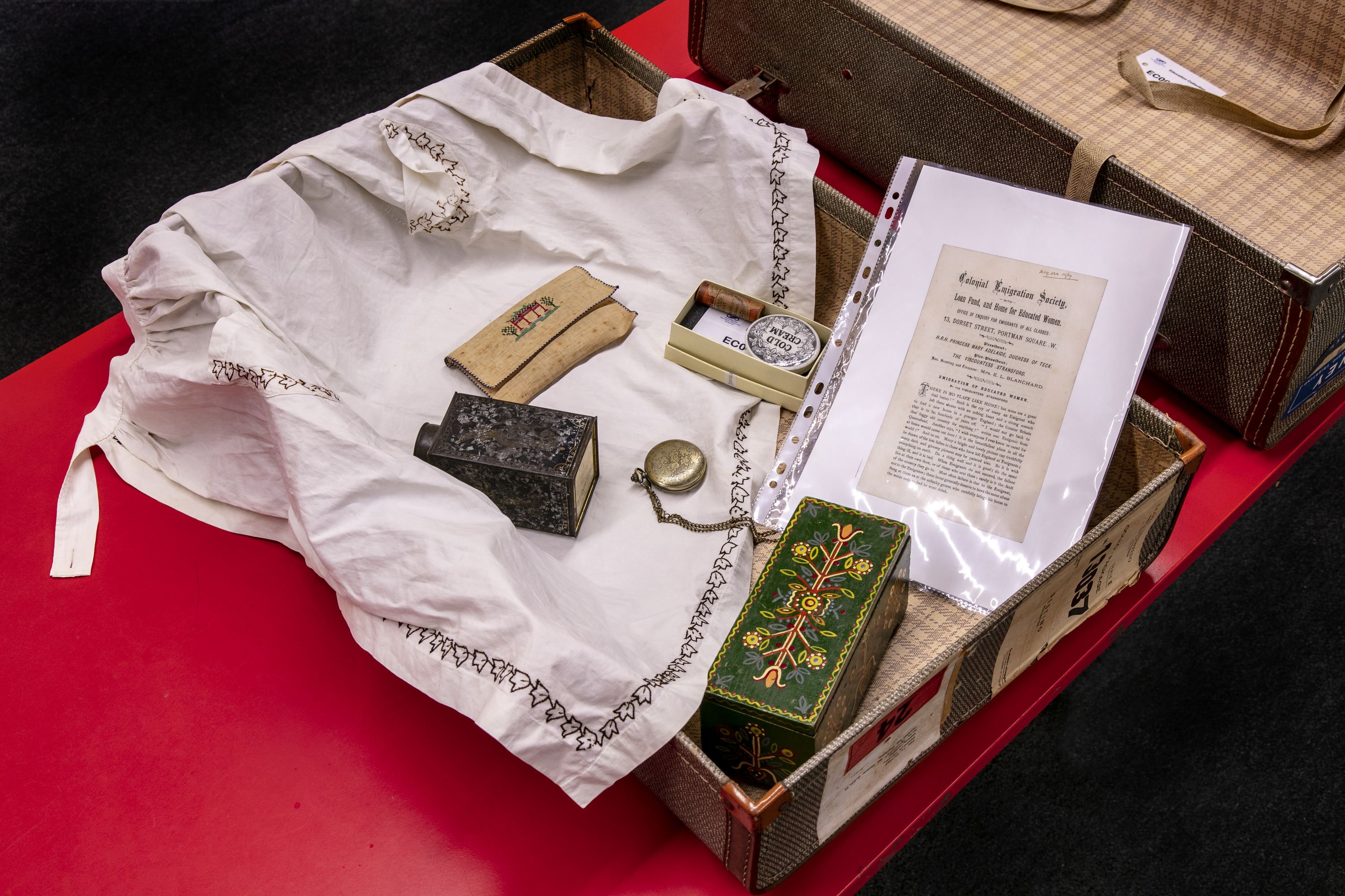 Photo showing objects arranged in travel suitcase. Objects include embroidered apron, sewing kit, metal container, fob watch, information booklet and decorated wooden box.