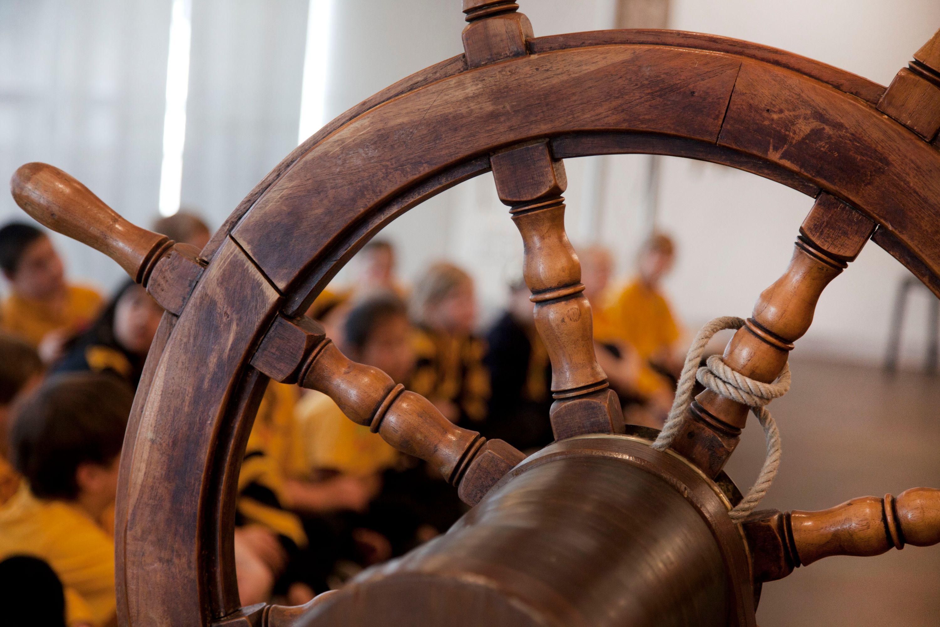 Photo focused on a ship's wheel tied with rope in the foreground with seated school children pictured in soft focus in the background.