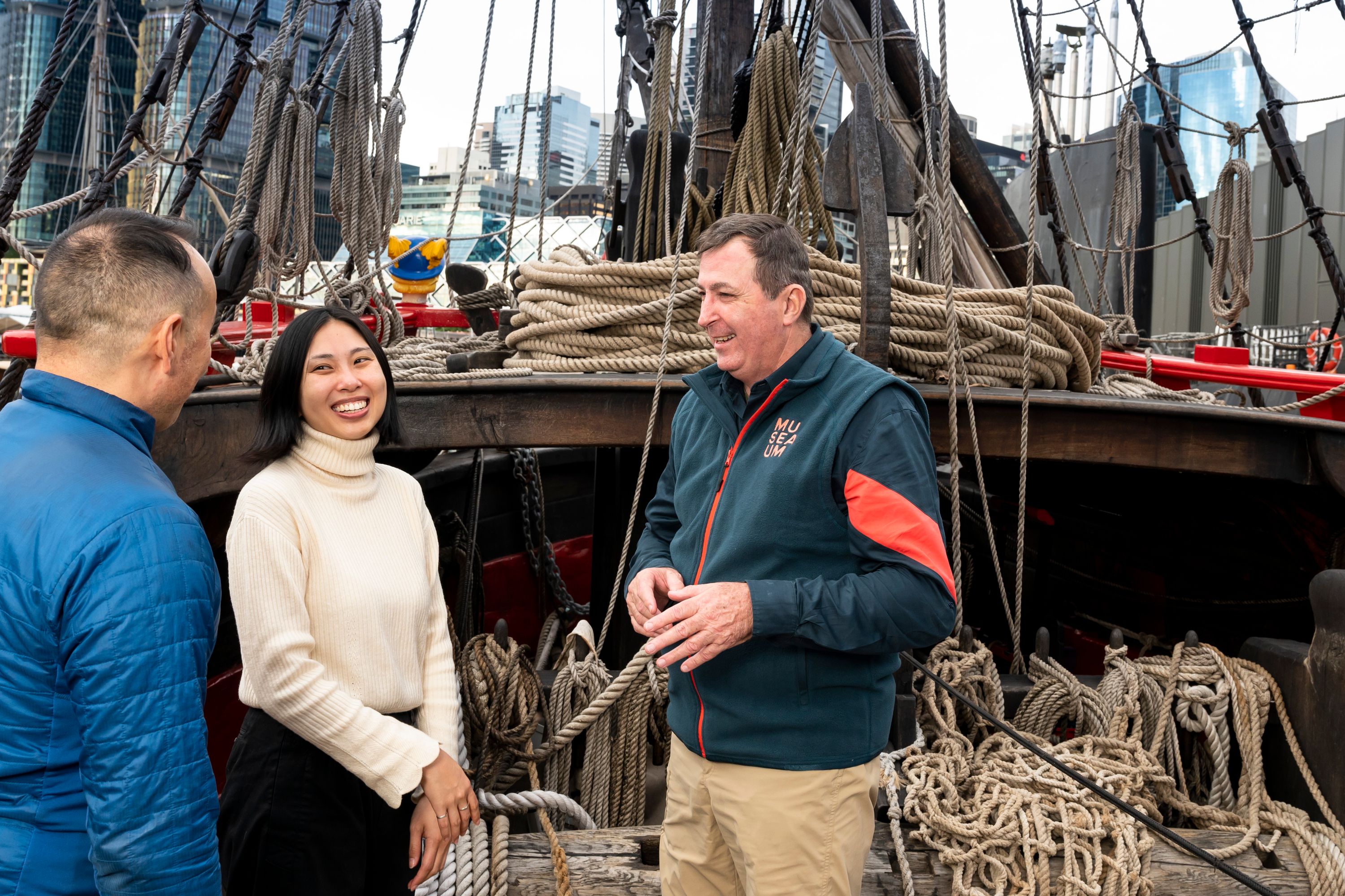 Photo of a male museum volunteer in uniform talking to a man and woman onboard a wooden boat.