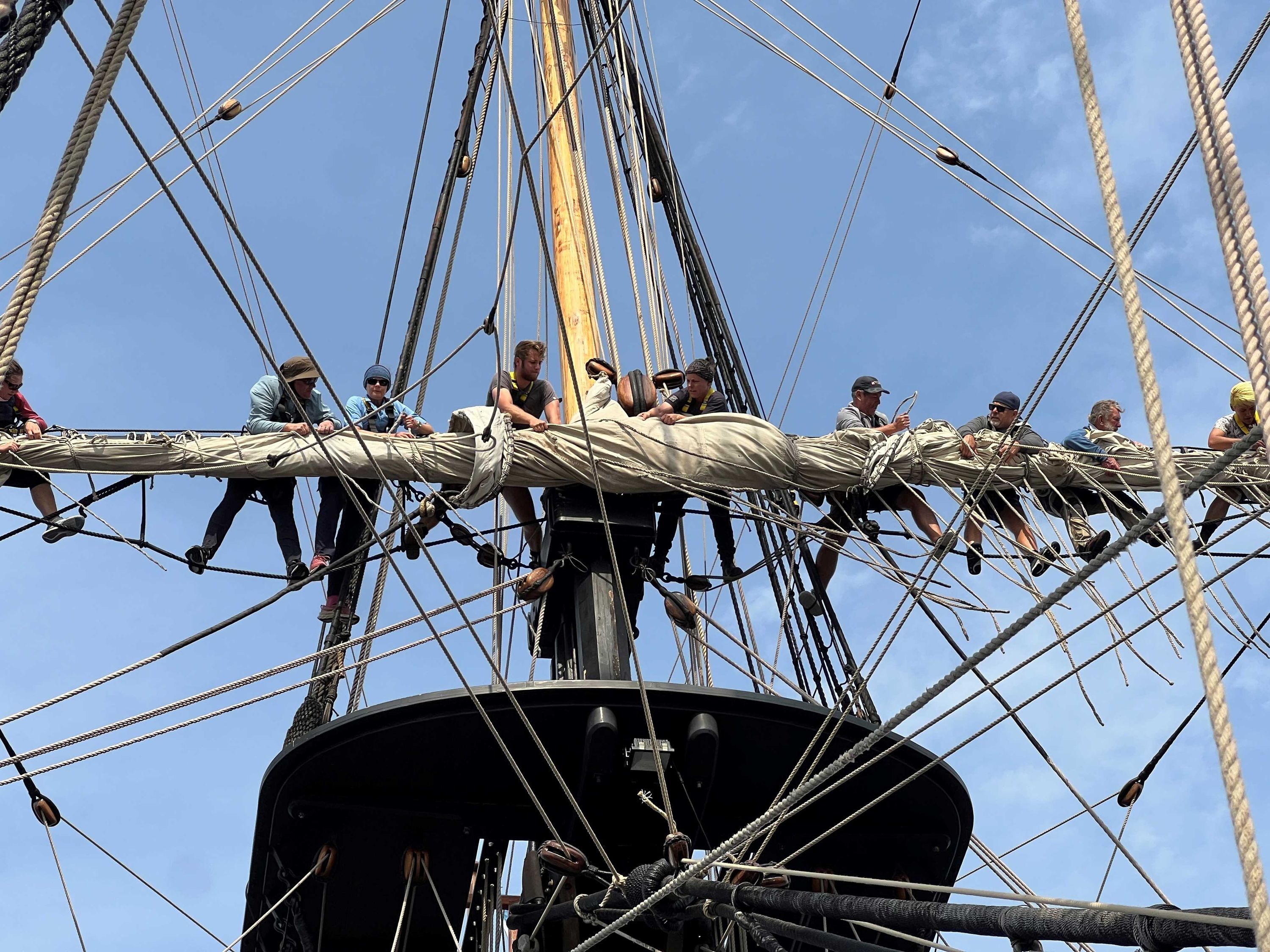 A photo taken of a tall ship's mast with people standing along the yard after having furled (packed away) the sail 