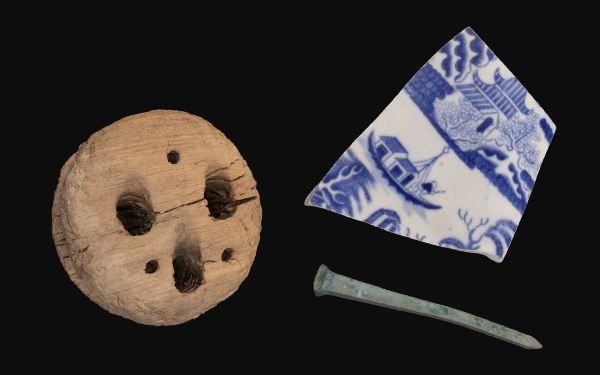 3 objects on a black background. A round wooden object, a broken piece of white and pottery and a green metal nail.