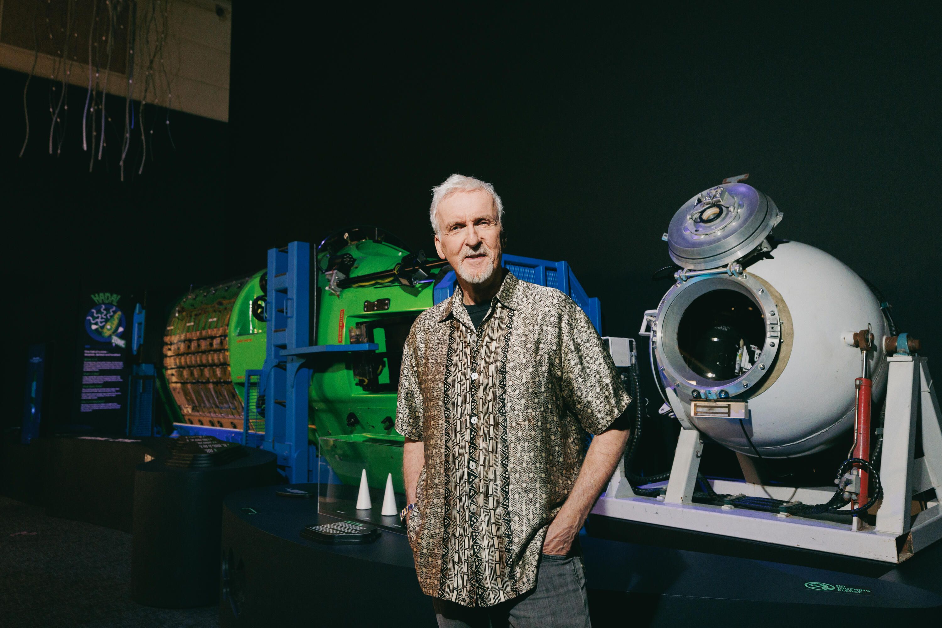 Photo showing a man standing in a museum exhibition in front of a large sphere and a large green/yellow submarine. He has grey hair and a beard, is wearing a light brown shirt with a subtle pattern on it, and has his hands in his pockets..
