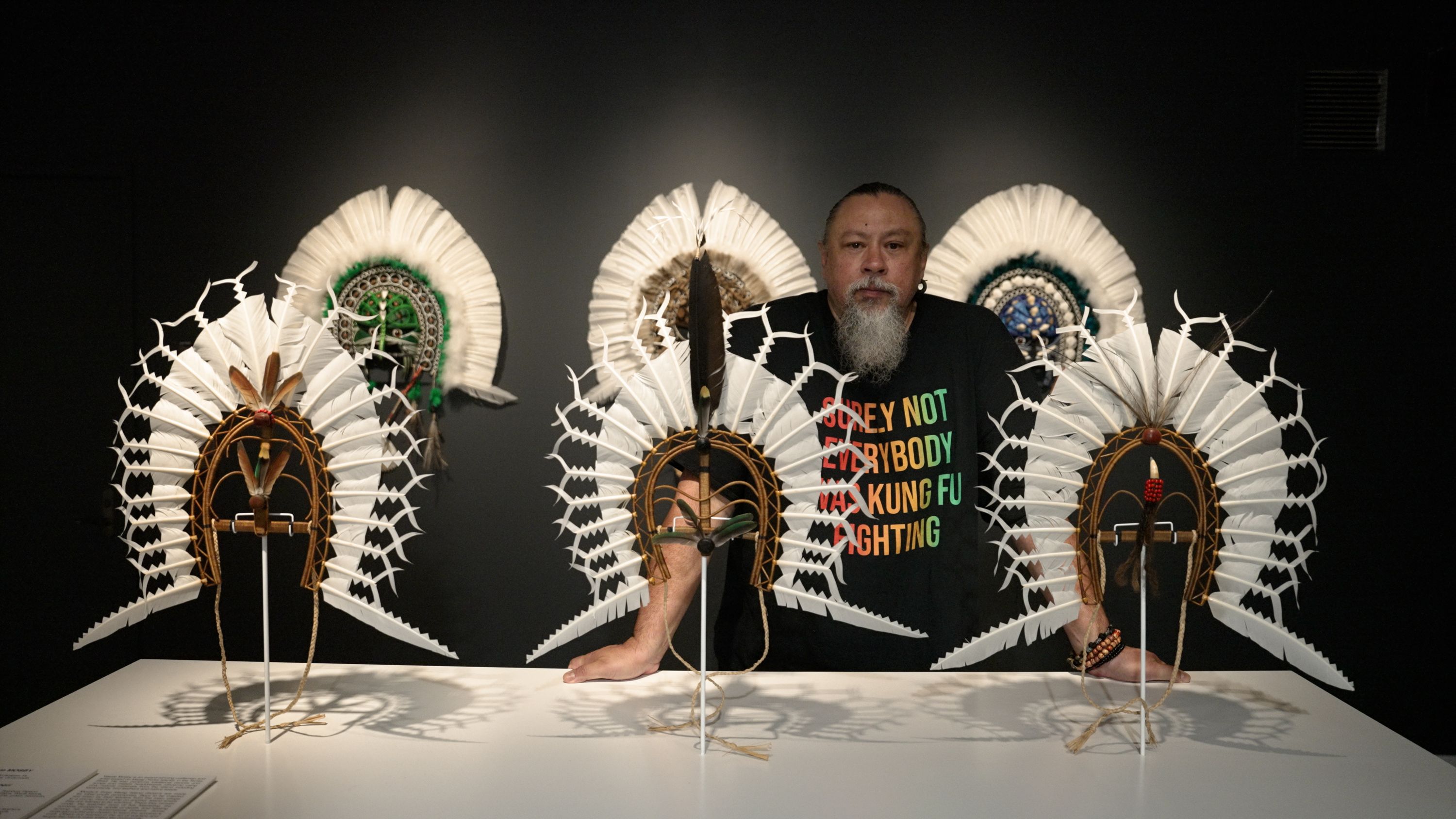 Photo of a man with a grey beard, wearing a black t-shirt and standing between headdress sculptures in a gallery.