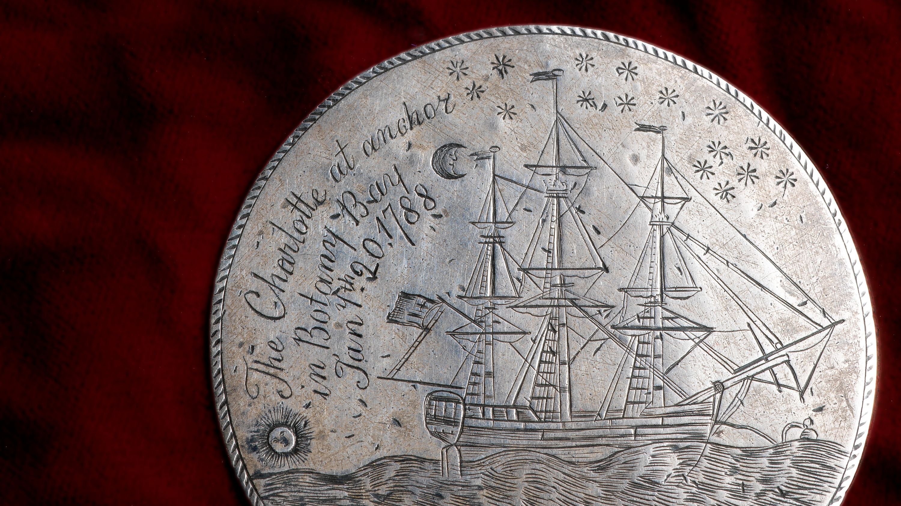 The CHARLOTTE Medal, a silver disc engraved on both sides. This photograph shows engraved a fully-rigged ship, the sun is near the horizon line on the lower left whilst a crescent moon and stars are inscribed on the upper right. Above are inscribed the words 'The CHARLOTTE at anchor / in Botany Bay / Jany. th 20, / 1788'.