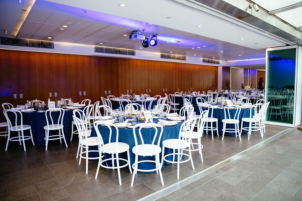 photo of event space with white chairs and large round tables with blue table cloths and place settings.