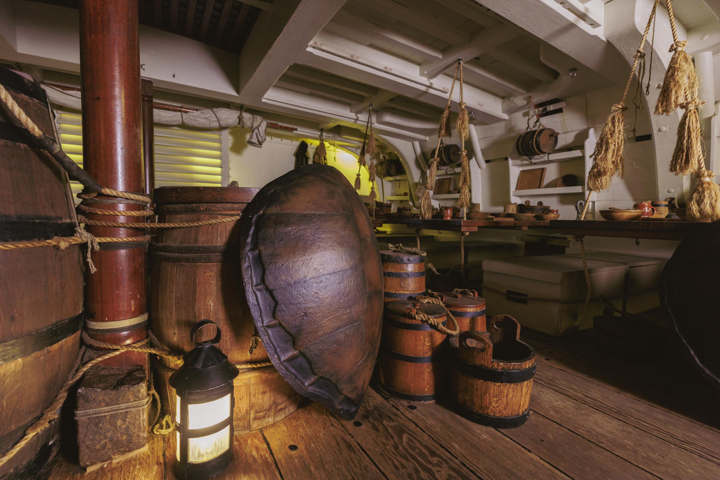 Photograph showing the below decks of a wooden ship with a large oval turtle shell standing up against large barrels.