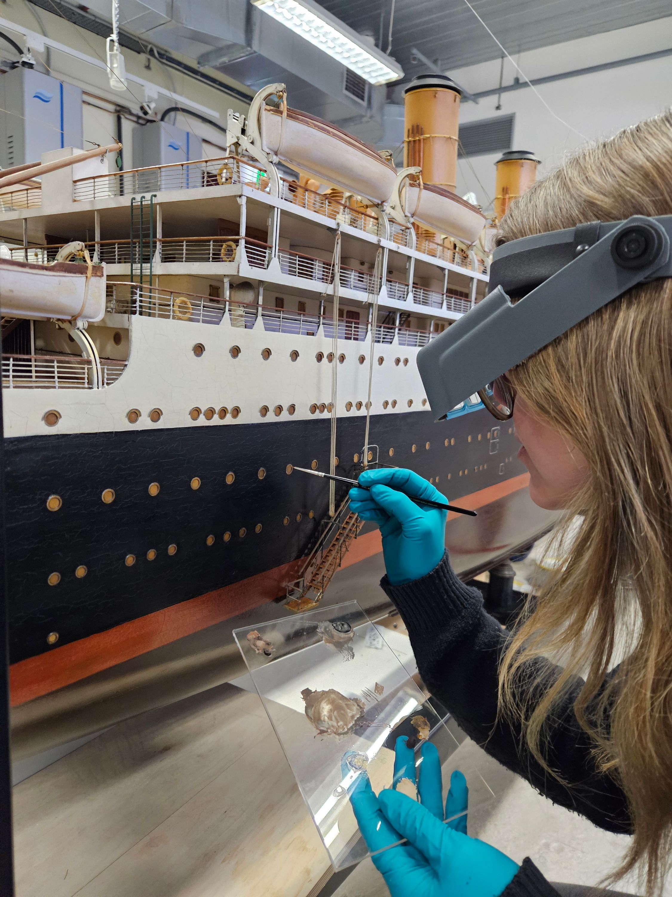 Photo showing a lady wearing magnifying glasses, using a small paintbrush to work on a large model of a cruise ship. 