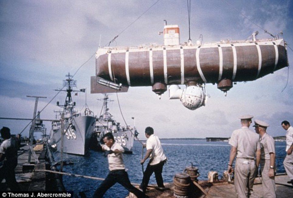 Colour photo of a submersible being lifted by a crane. There are men in the foreground an Navy vessels behind.