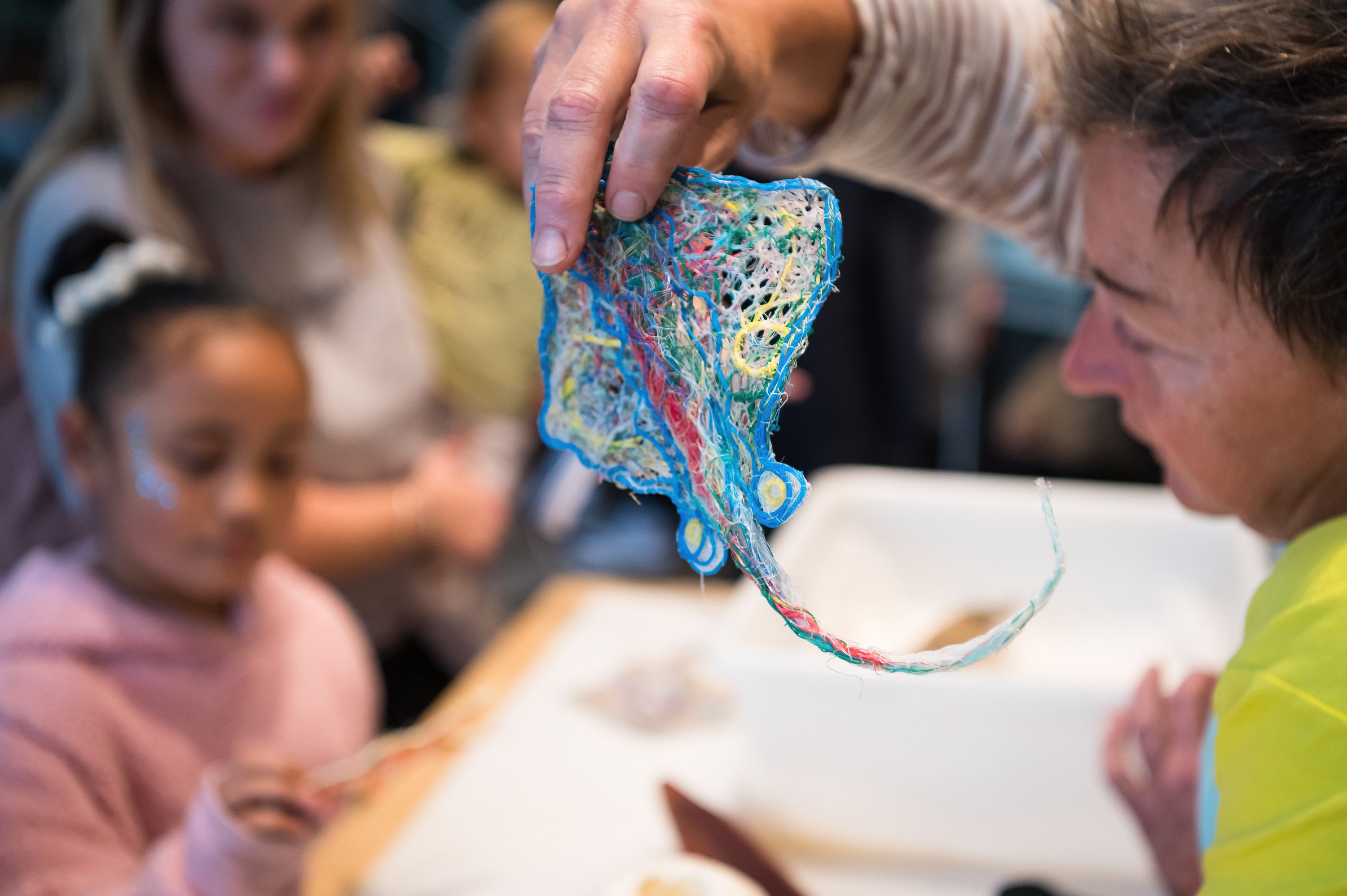Photo of a woman holding a sculpture of a small stingray made from recycled fishing line, with children in the background. 