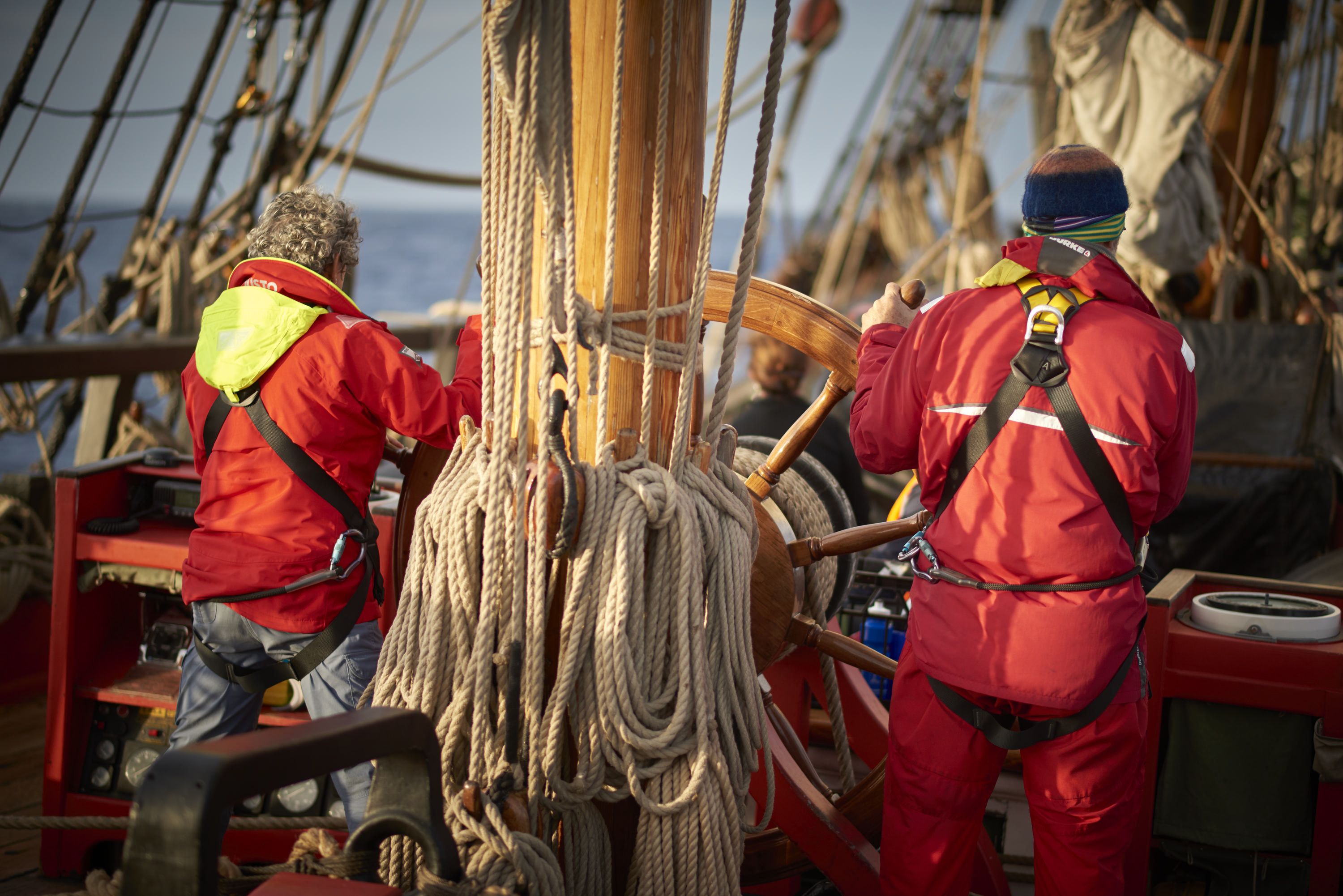 Above deck, voyage crew at the ship's wheel, dressed in wet weather gear. 