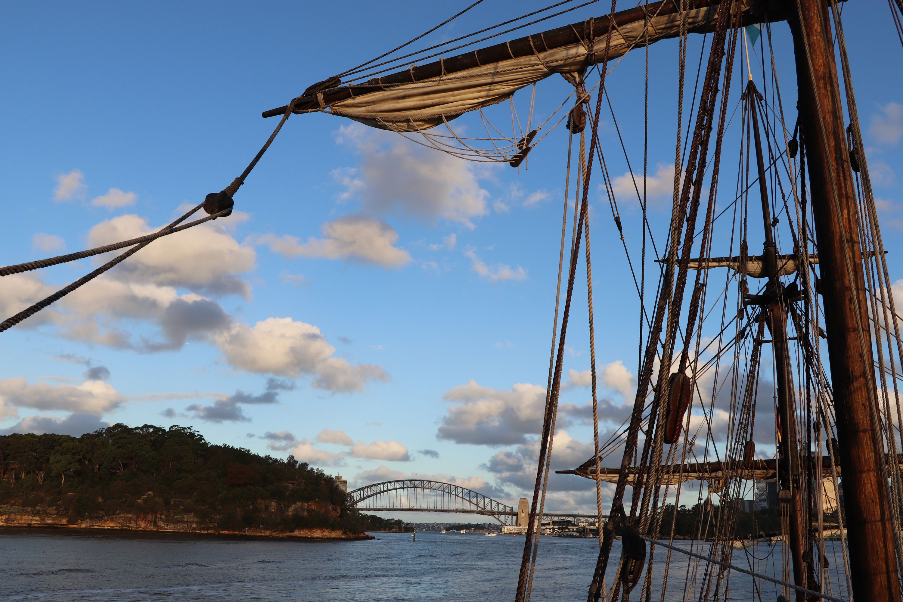 Photo taken from a tall ship with the mast in the forground, looking at the Sydney Harbour Bridge in the background and a blue sky with white clouds