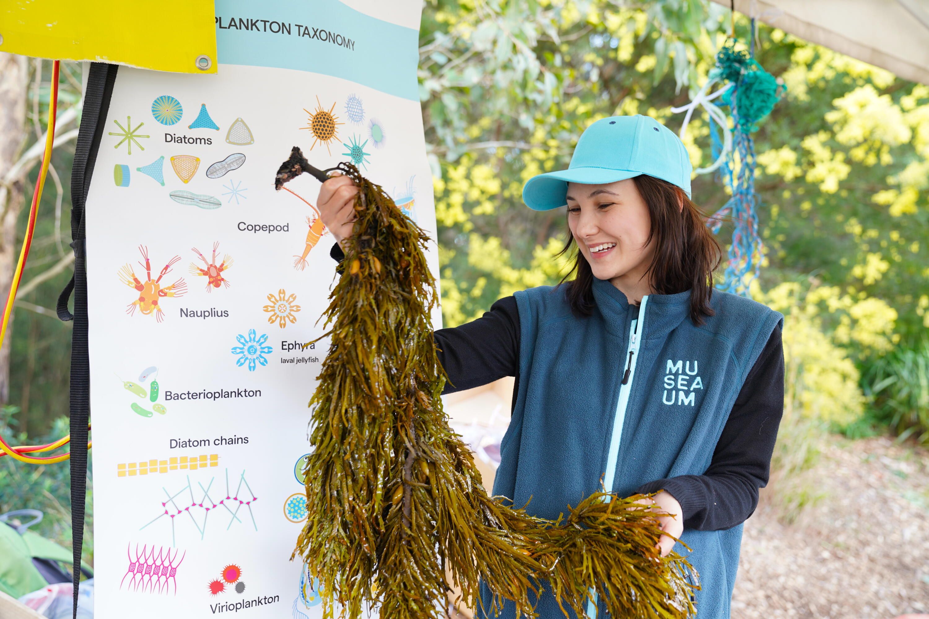 Photo of a woman wearing a museum uniform vest and hat holding up a large bunch of seaweed