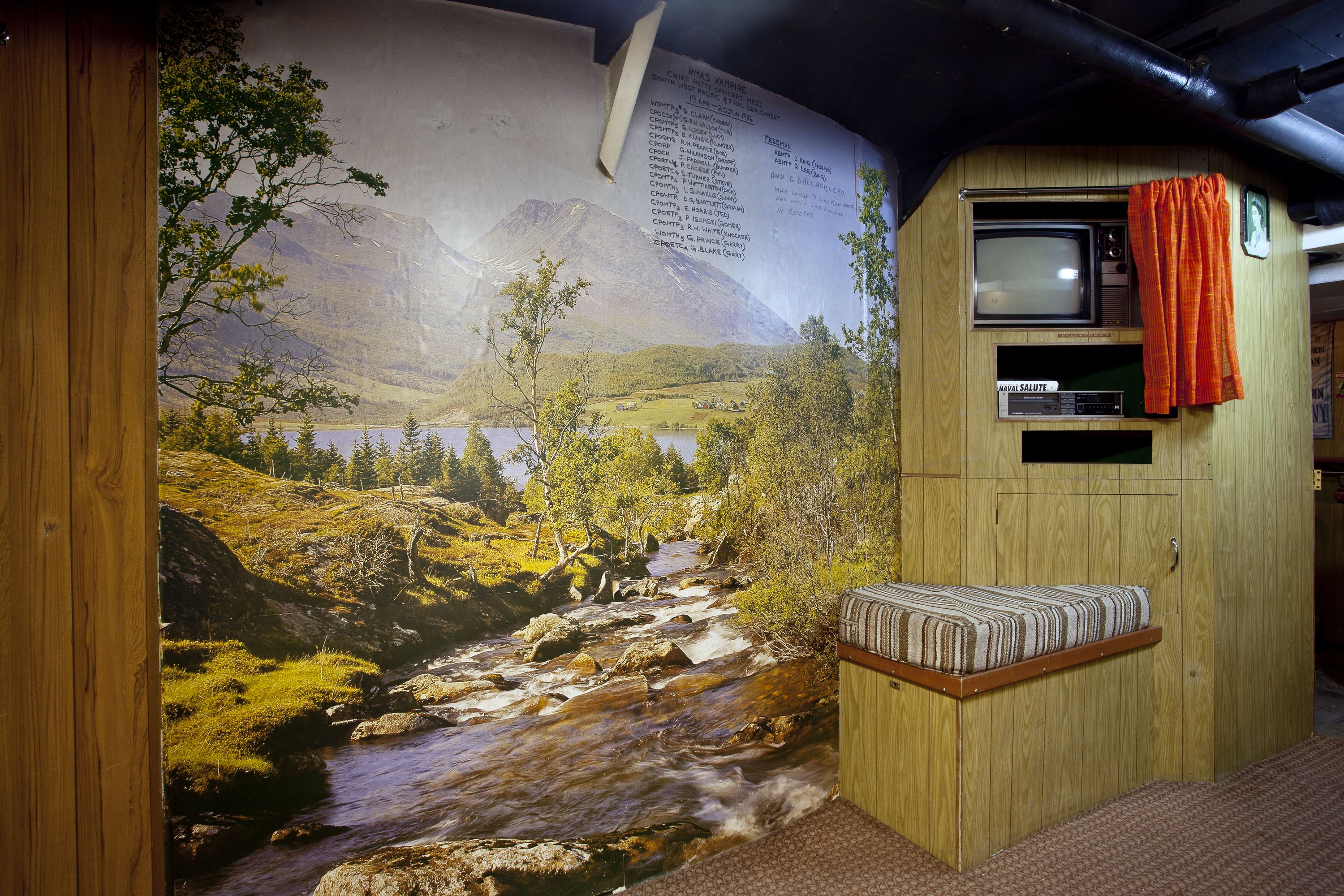 Photograph of inside a navy ship, a room decorated with a scenic photographic landscape wallpaper.  on the right is a TV screen and VHS video recorder inside a wood panelled entertainment unit.