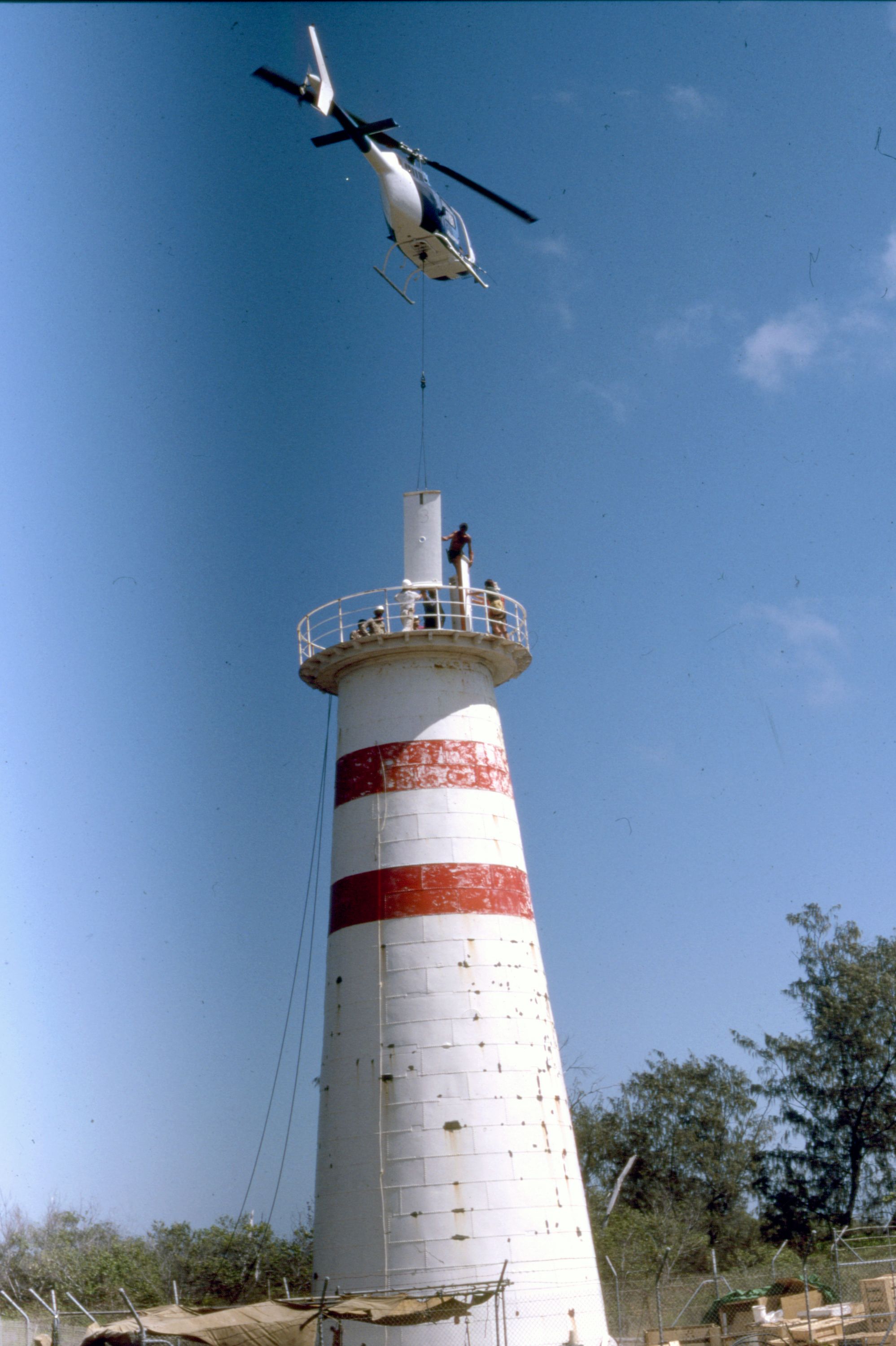 Photo showing the dismantling of a lighthouse with a helicopter flying overhead caring a metal sheet.