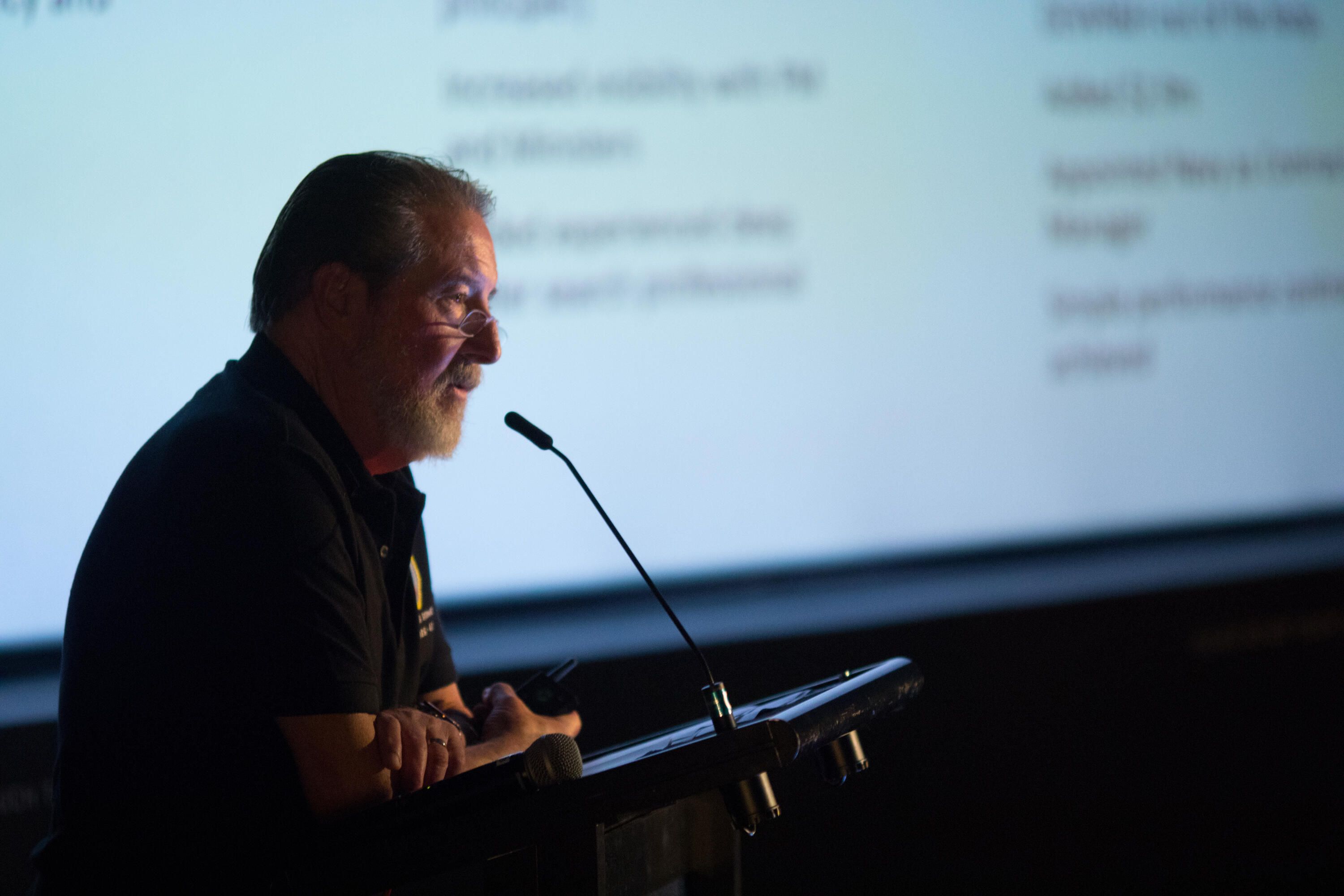 Photo of a side view of a male speaker taking into a lectern microphone, with a presentation in the background.