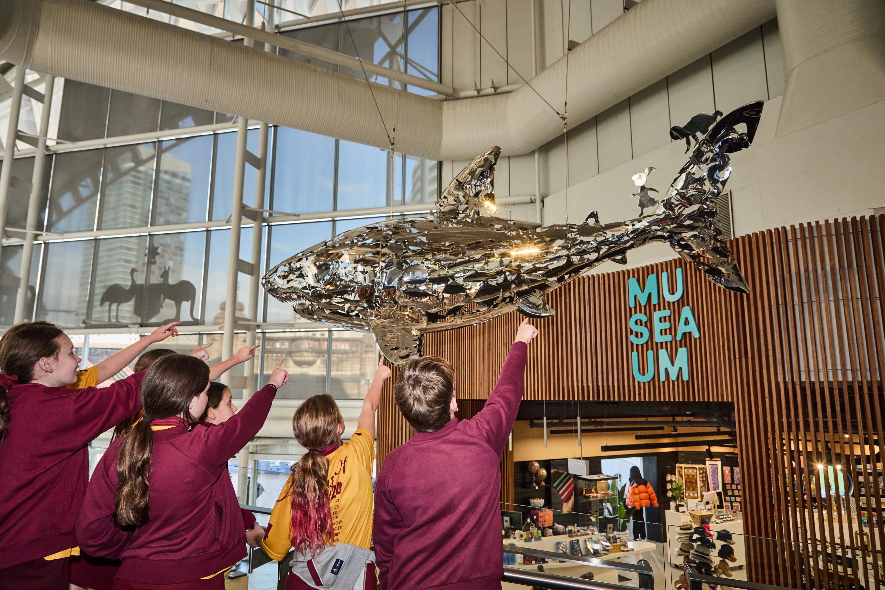Photo taken inside the museum, with a group of students pointing up at a silver sculpture of a shark.