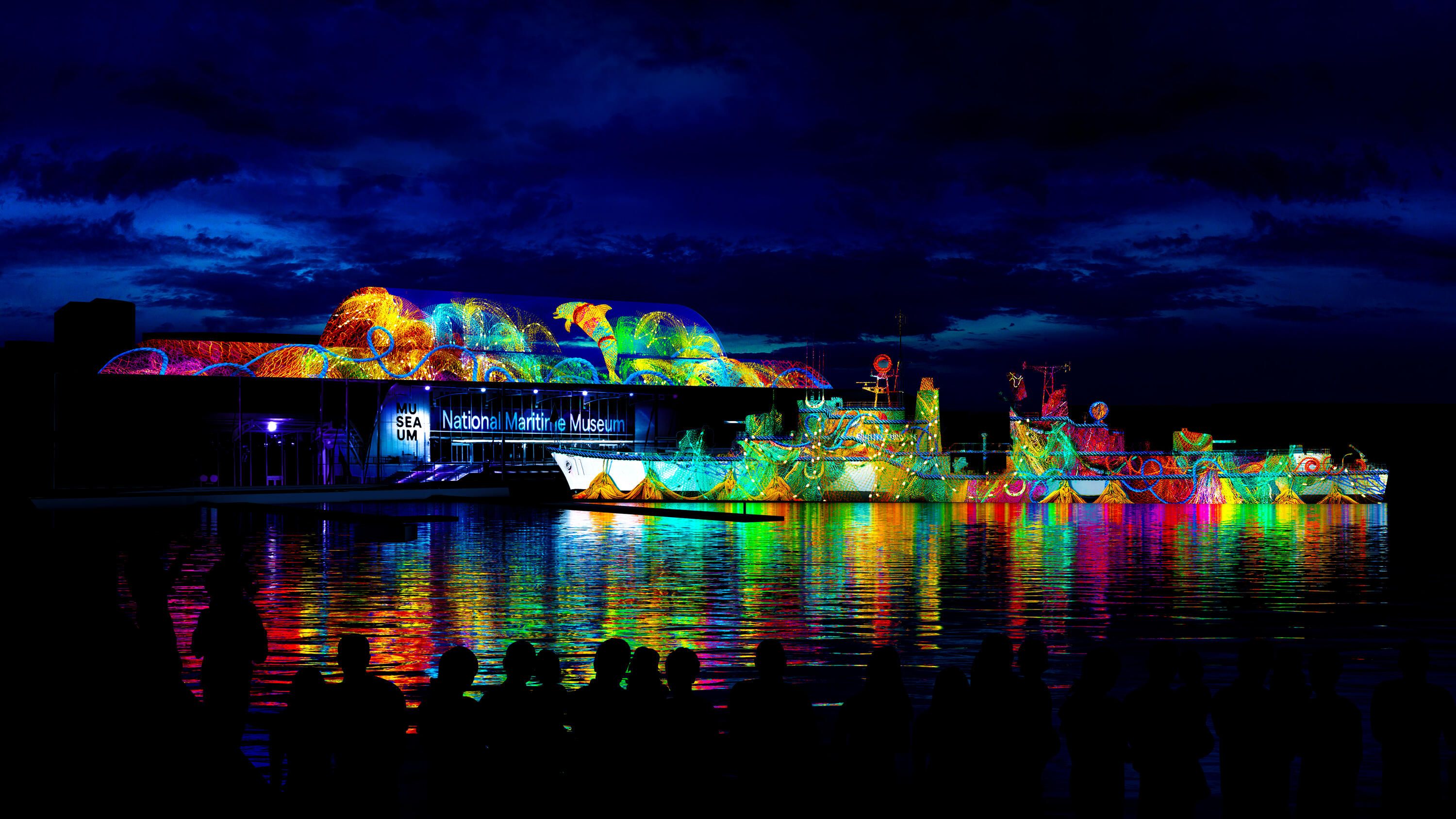 photo of colourful projection on the side of the museum and navy destroyer at night, with multicoloured reflections on the water. 