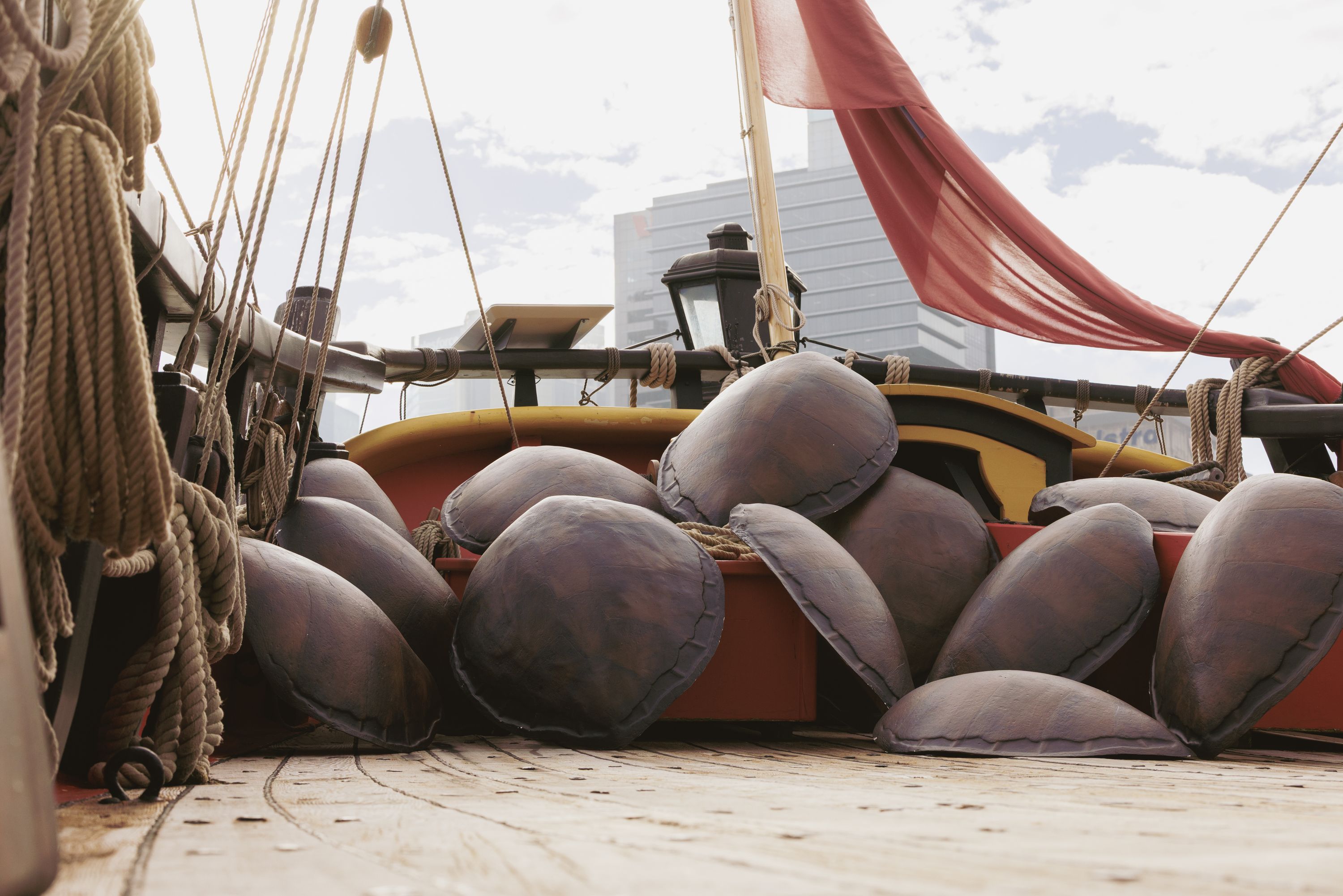 Photo showing the back of a wooden tall ship with a collection of turtle shells on the deck. There is a red flag flying on the ship, and city buildings in the background.