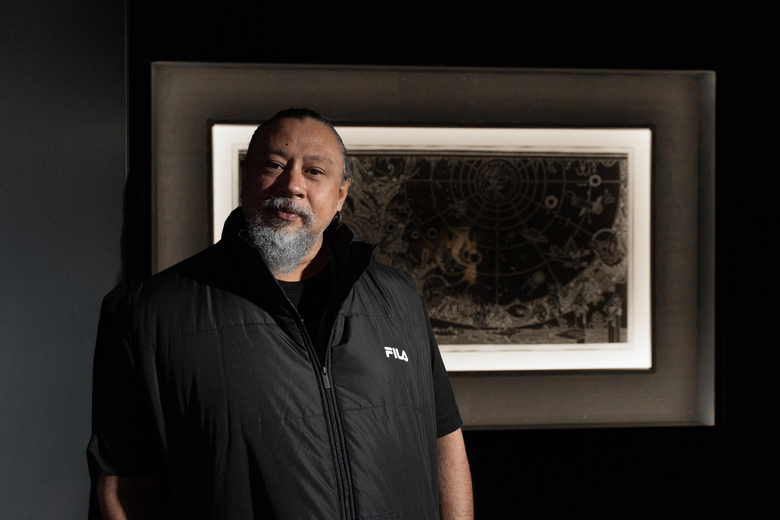 Photo of a man with a grey beard, wearing a black vest and shirt. He is standing in front of an artwork in a dark gallery space. 