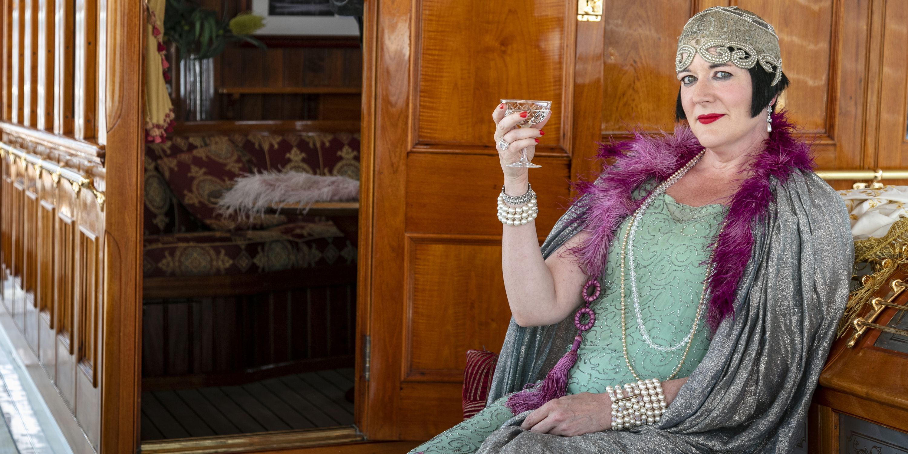 A lady poses in vintage 1920s flapper style dress, shawl and hat with a glass.