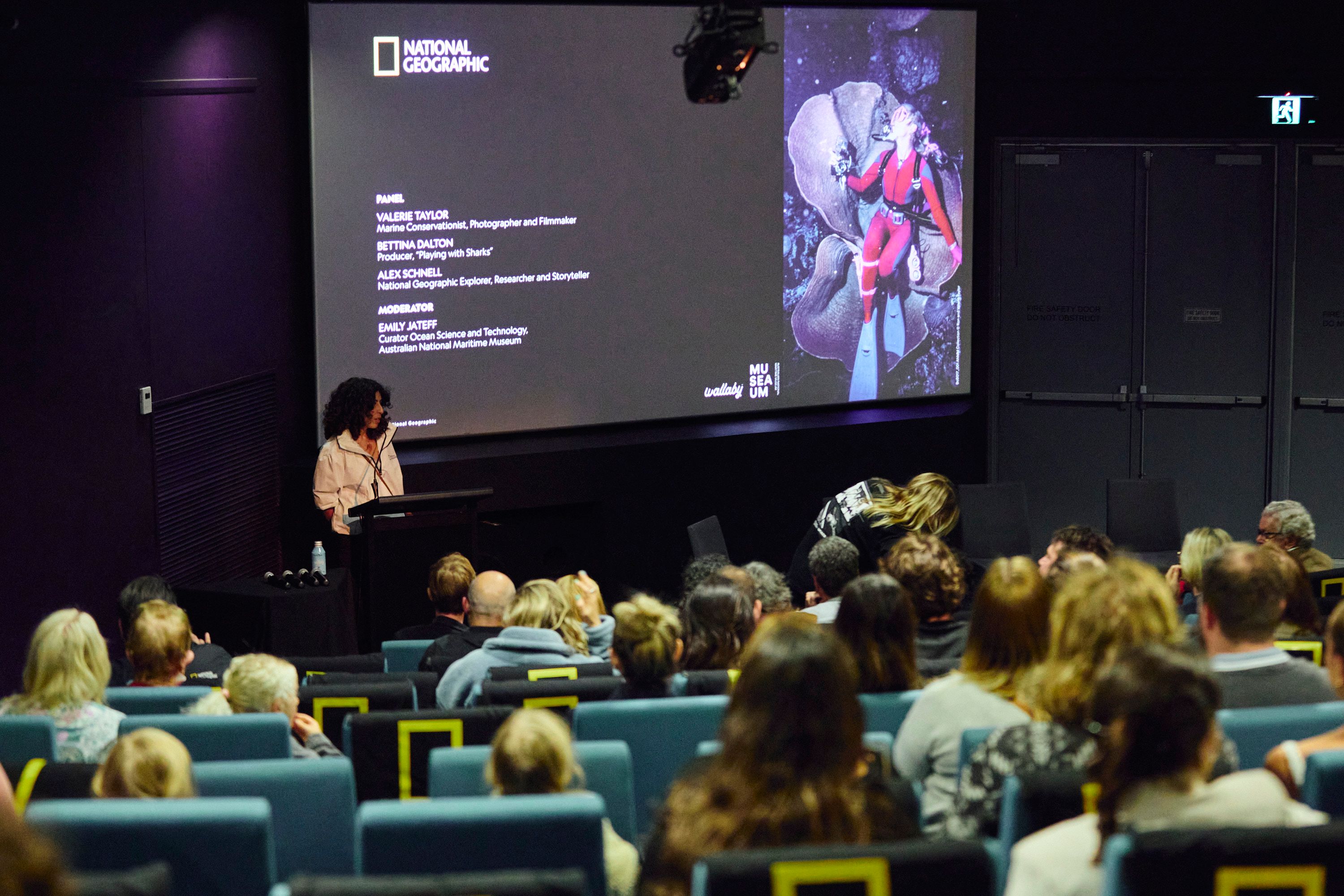 Photo of people sitting in a theater with a woman presenting at the front.