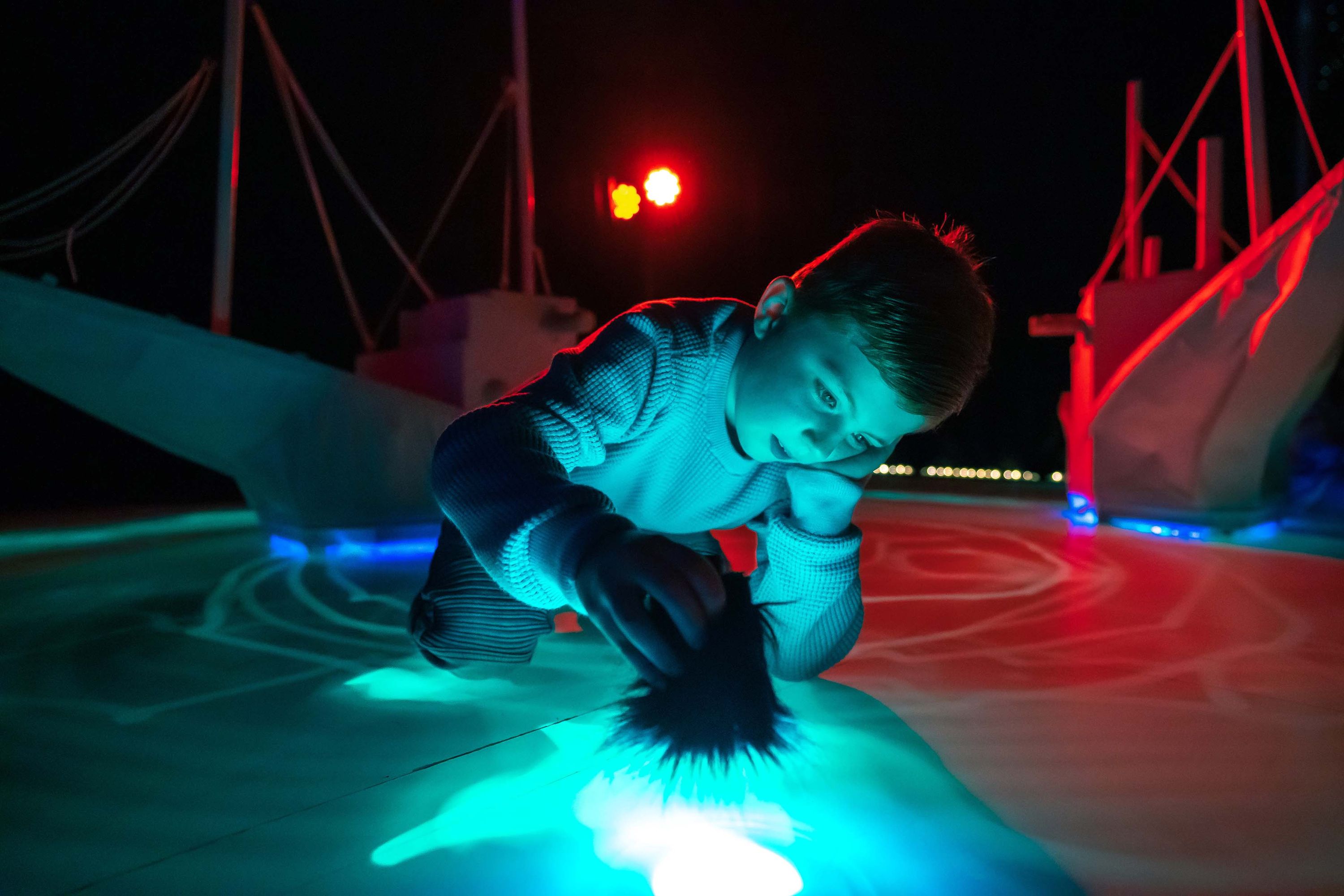Photo showing a boy in a dark space with neon lighting, crawling on the floor shining a torch.  