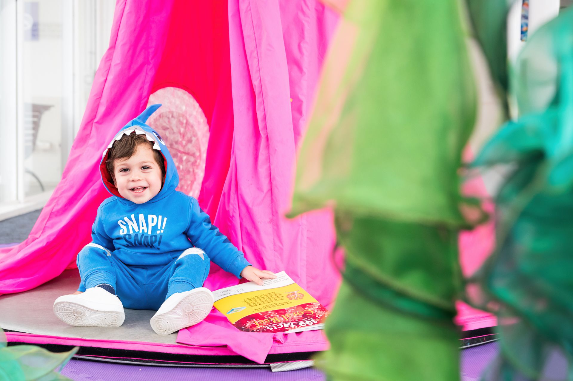 Photo of a seated child in a shark hoodie, smiling in the direction of the camera.