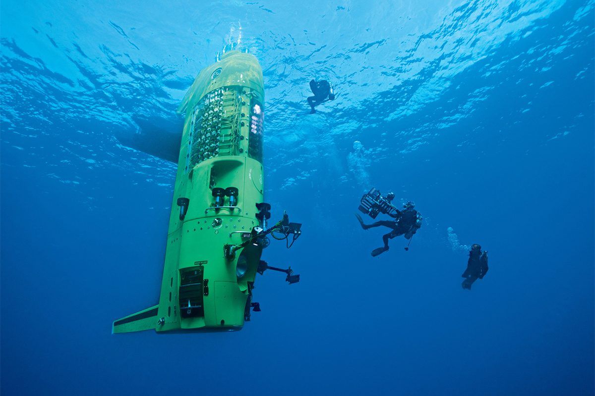 Underwater photo looking up at a tall yellow/green submarine near the surface of the water with several divers around it.