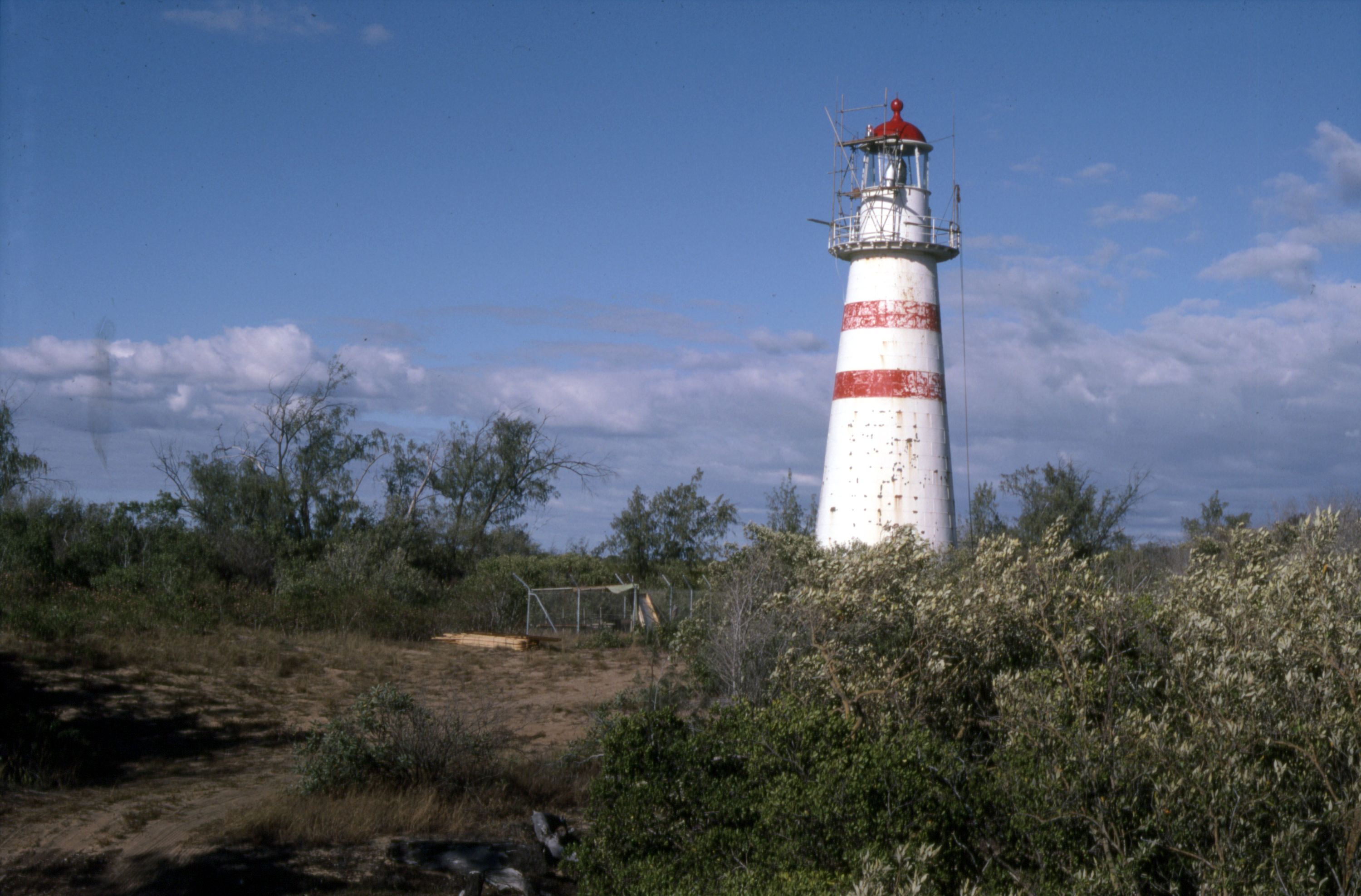 Photograph of a white lighthouse with 2 red stripes and a red roof, surrounded by small shrubs and trees.