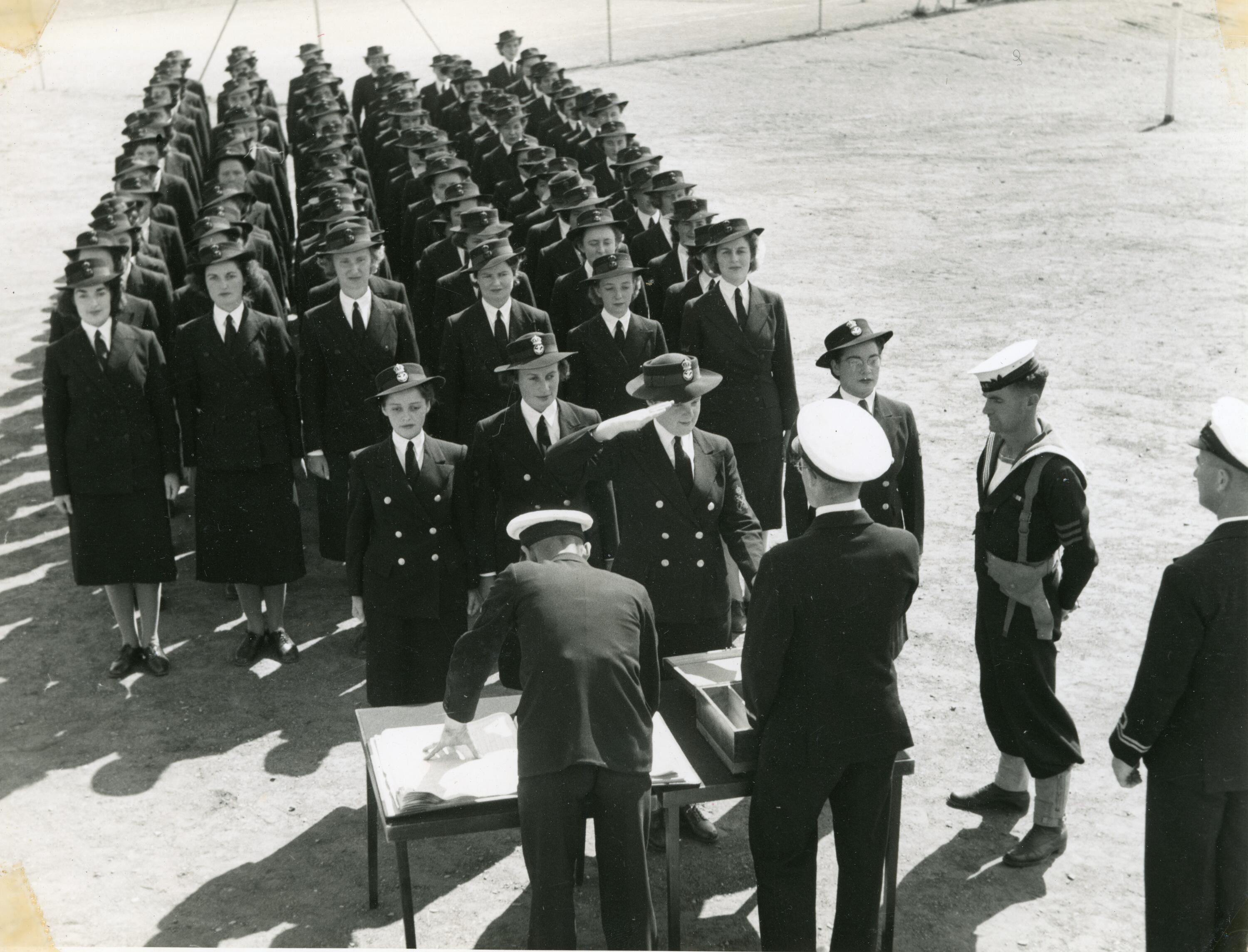 A photo showing rows of women in uniform standing in military formation, with 3 women in the front standing at at a table with male officers. 