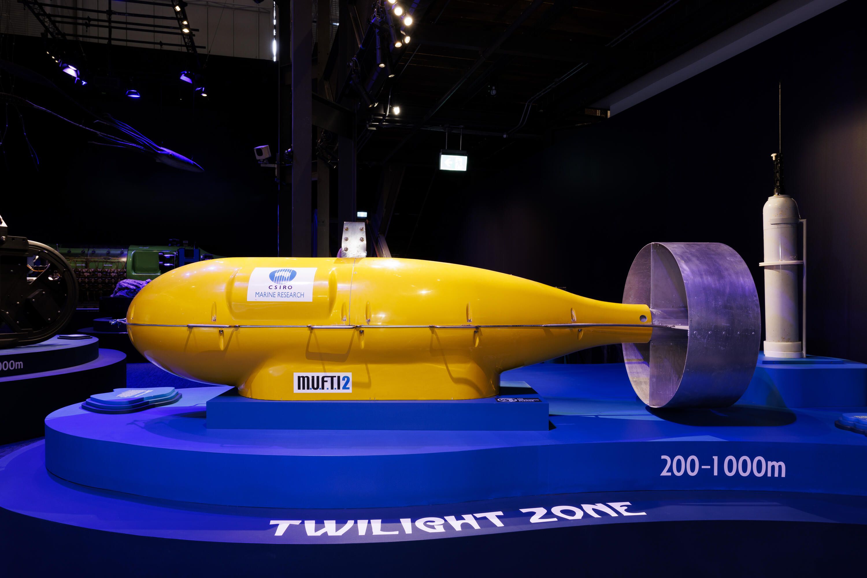 Photogrtaph of a large, bright yellow, torpedo shaped object, in display in a museum exhibition.
