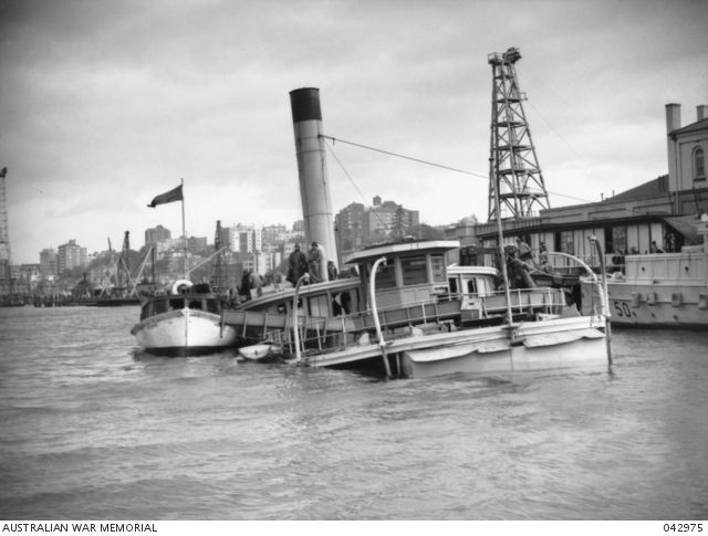 Black and white photo showing the partly submerged wreckage of a ship, it's top deck and funnel sticking out of the water at an angle.