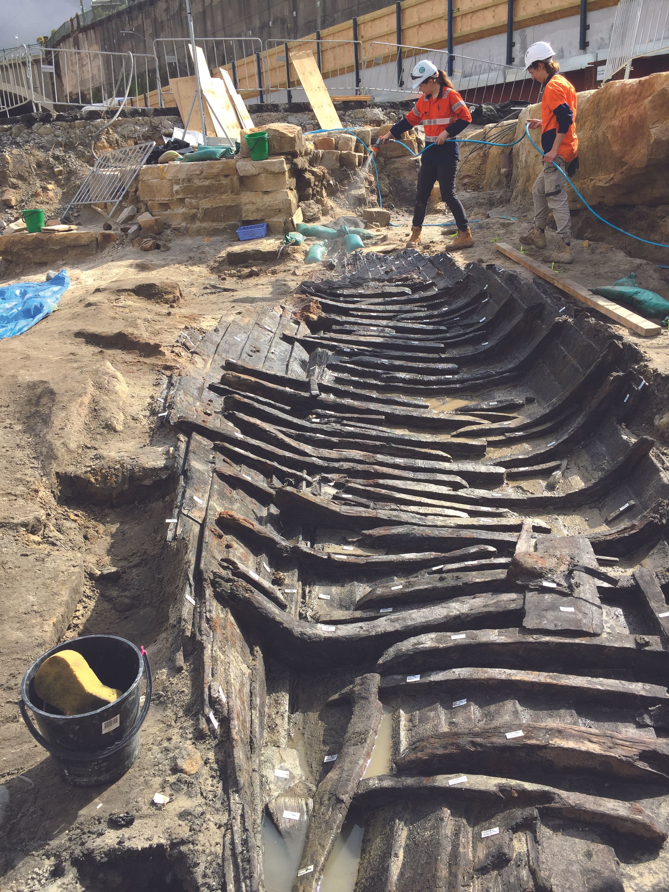 Photo showing an archaeological dig, with 2 people wearing fluro shirts and hard-hats using a hose to wet a wooden boat in the ground 