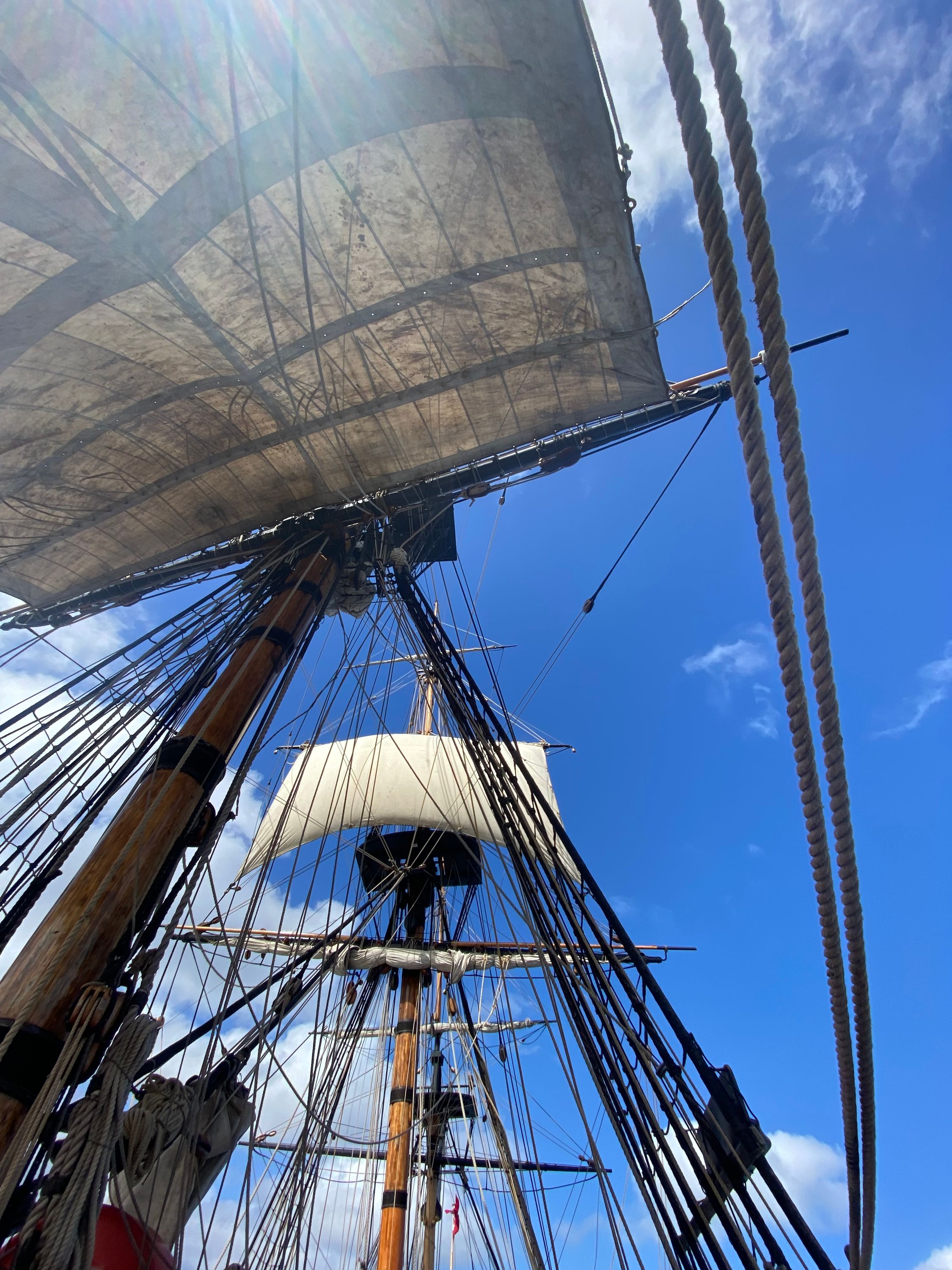 Photo taken looking up at the sails and rigging of a tall ship. The blue sky and white fluffy colours are above.