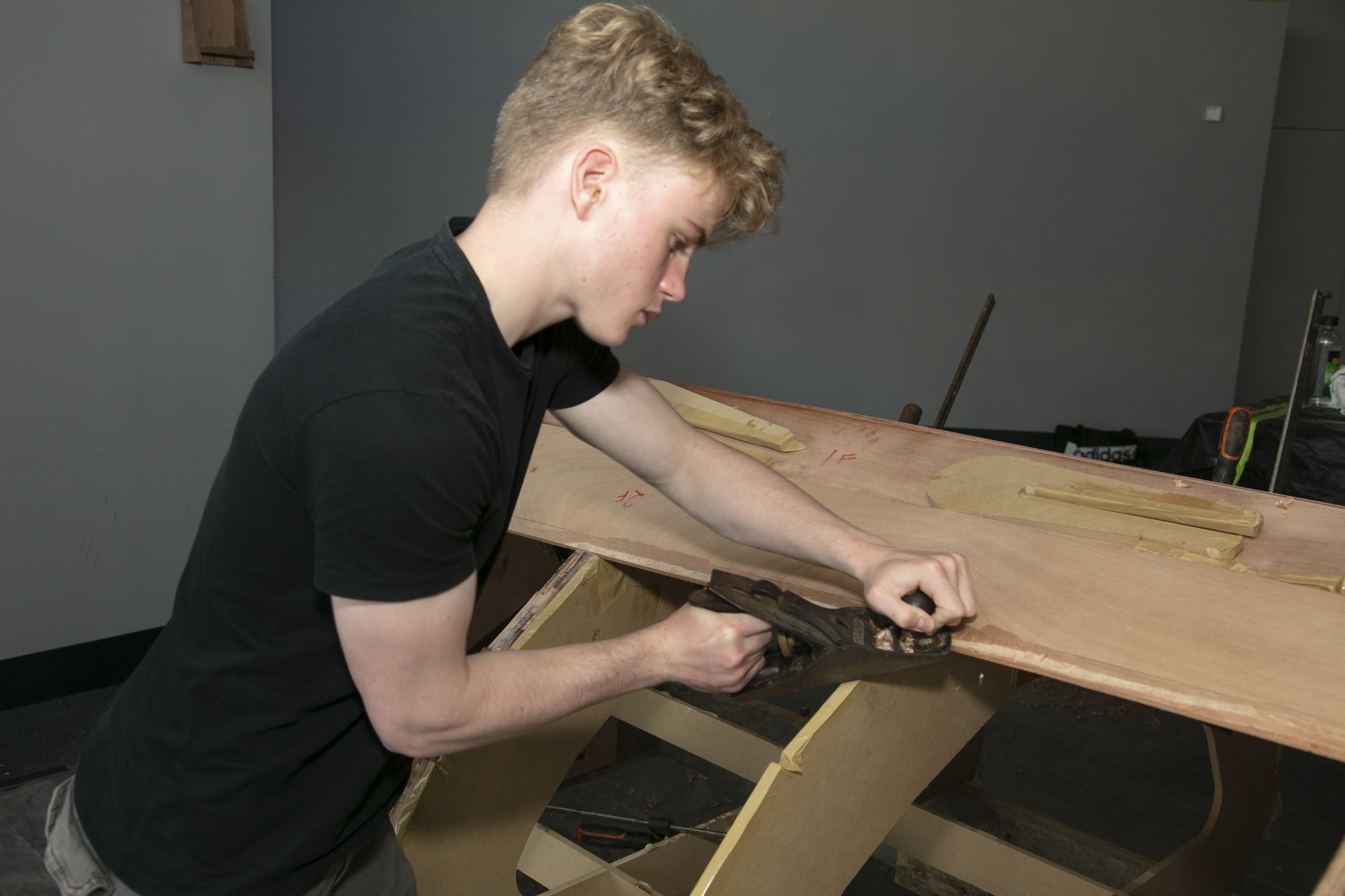 A teen boy in a black t-shirt is using a hand tool to build a wooden boat.
