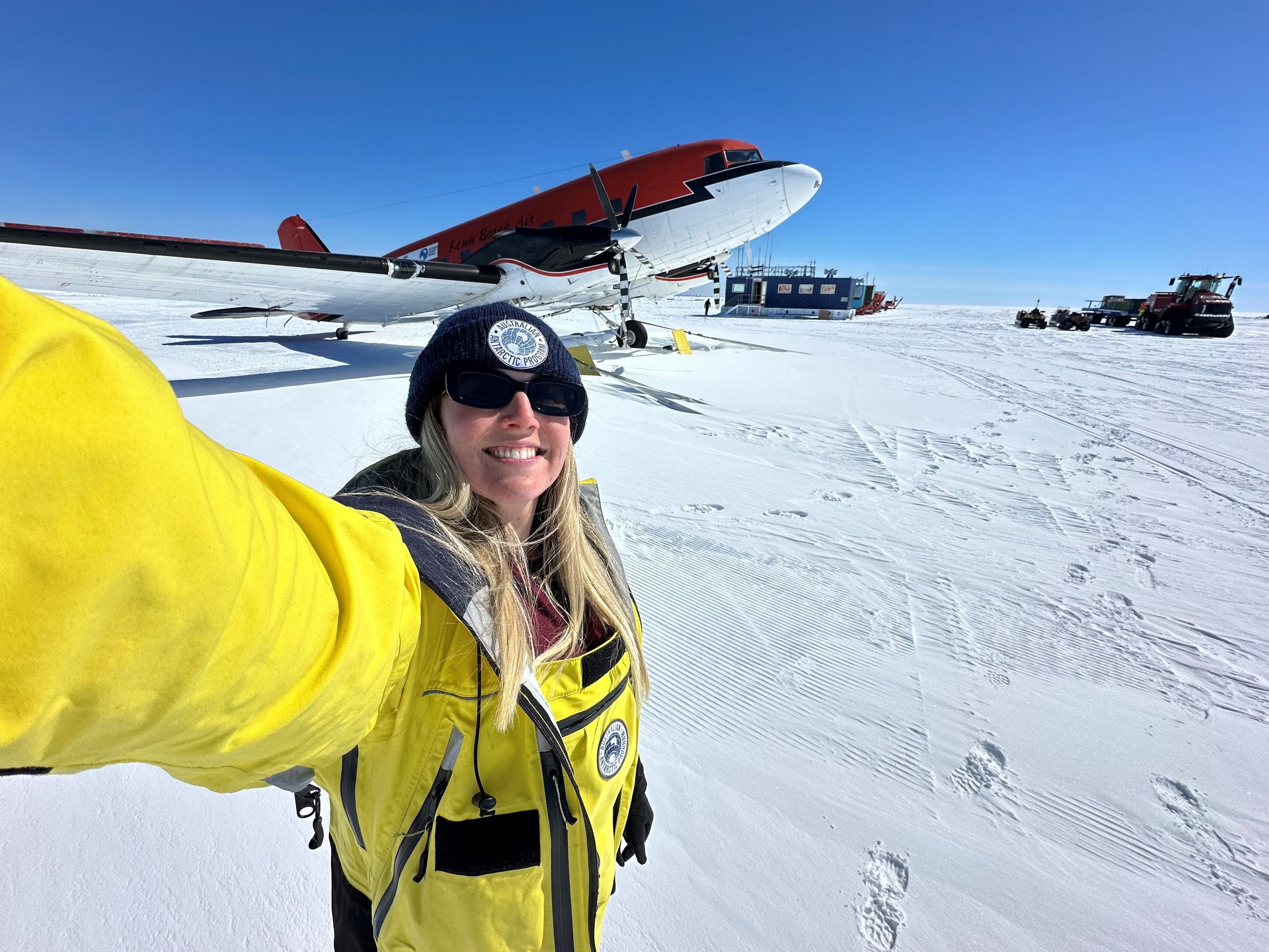Photo of a woman with long blonde hair, warm clothes, beanie and sunglasses taking a selfie in a snowy landscape with blue sky and a plane in the background.