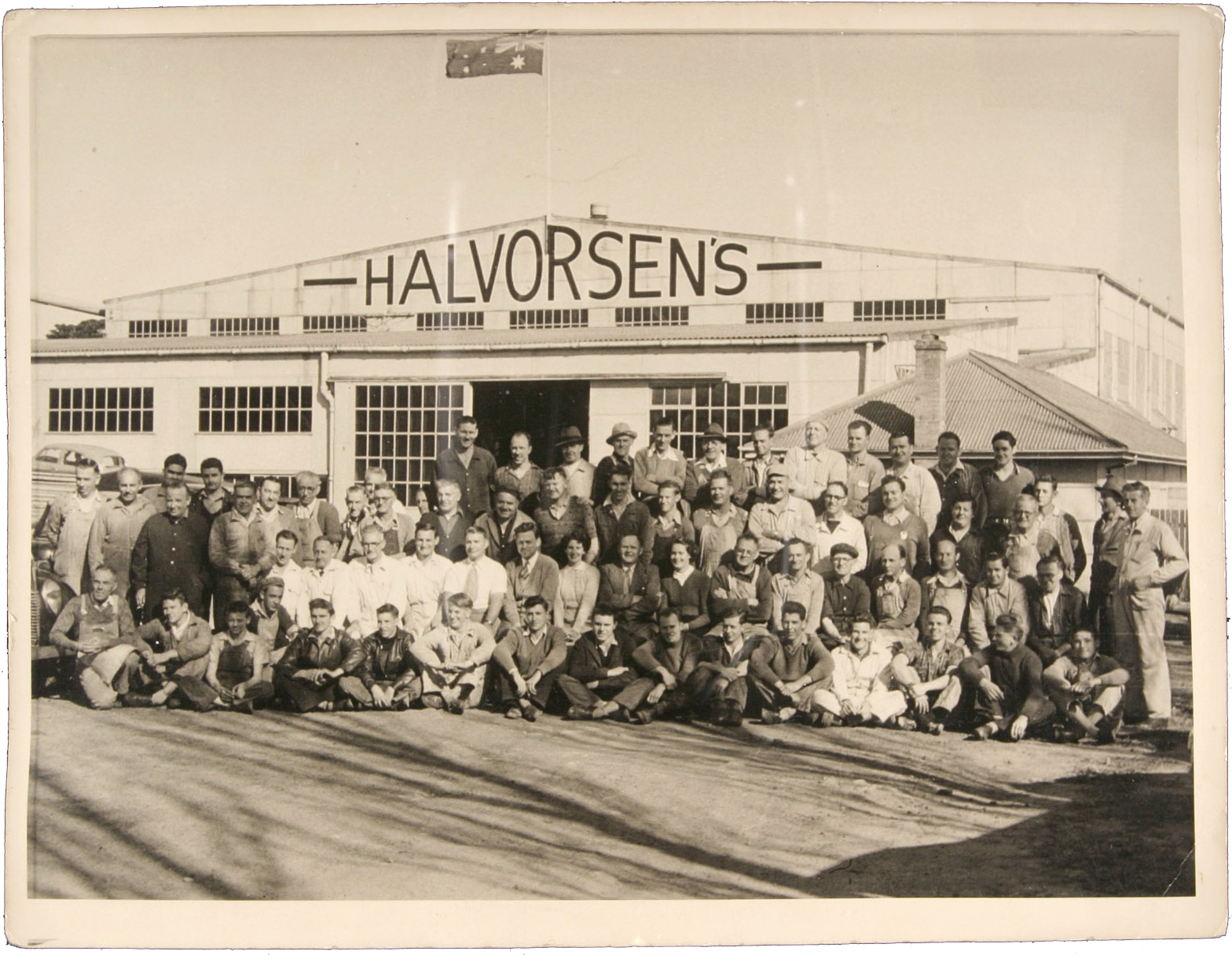 Black and White photograph showing a large group of boat builders, managerial and administration staff employed at the Halvorsen boatyard.  There is a large factory building with "Halvorsens" painted on it, in the background. 