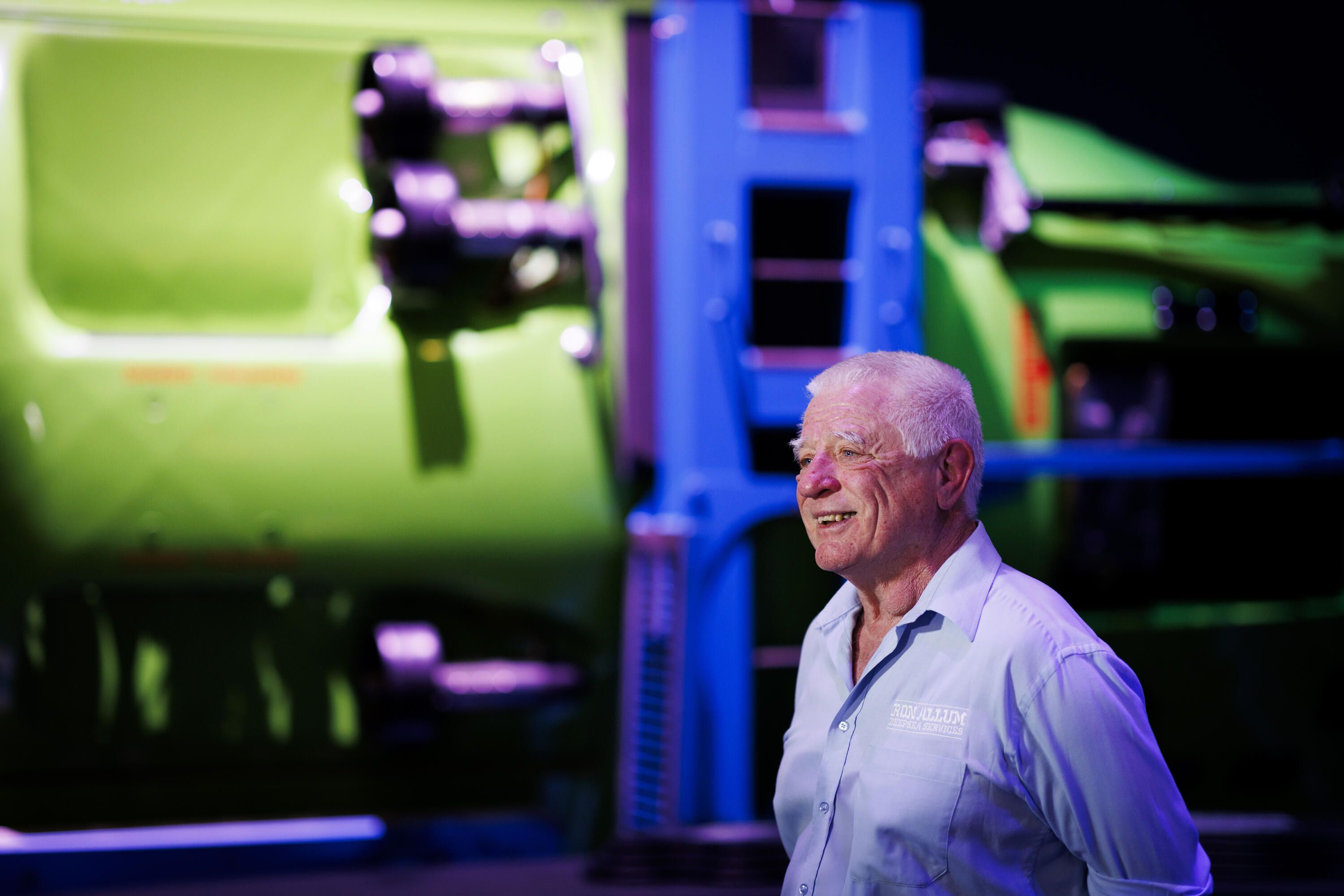Photo showing an older man with short white hair, wearing a light blue button up shirt standing in a museum exhibition in front of a submarine.