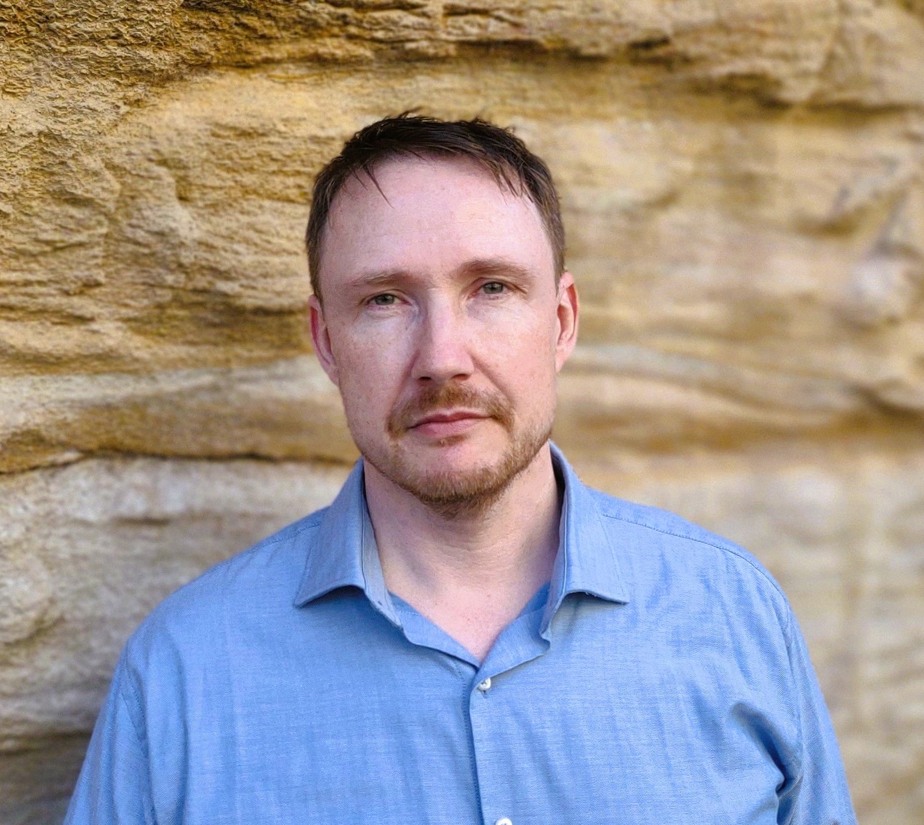 Photograph of a man with light skin, short hair and a blue shirt standing in front of a sandstone wall.