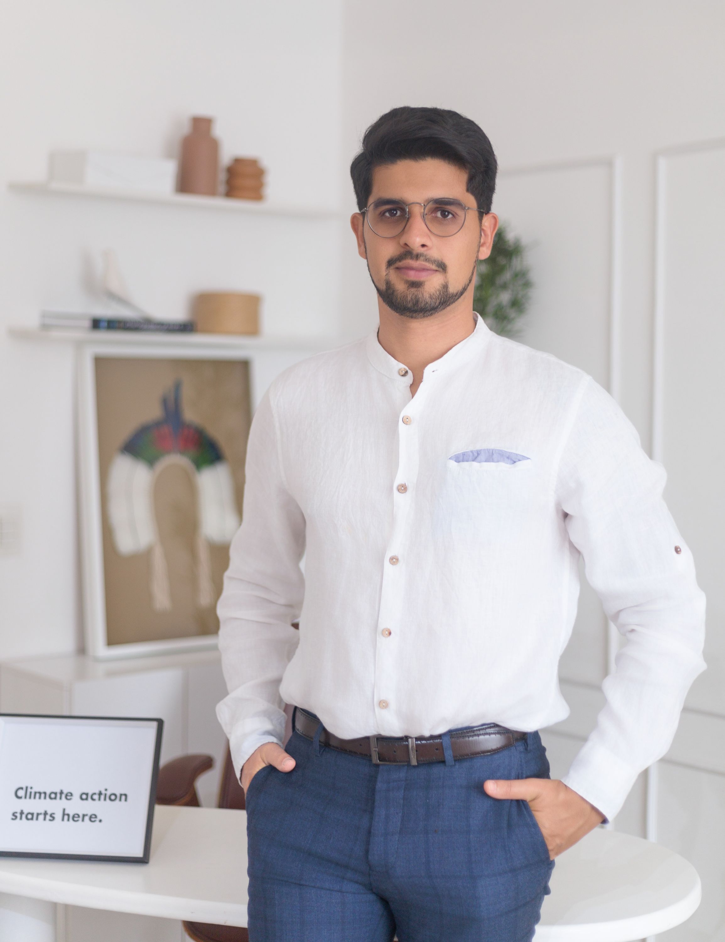Photo of a man wearing a white button up shirt and blue pants starting inside a room white white walls and a small amount of decoration in neutral colours.