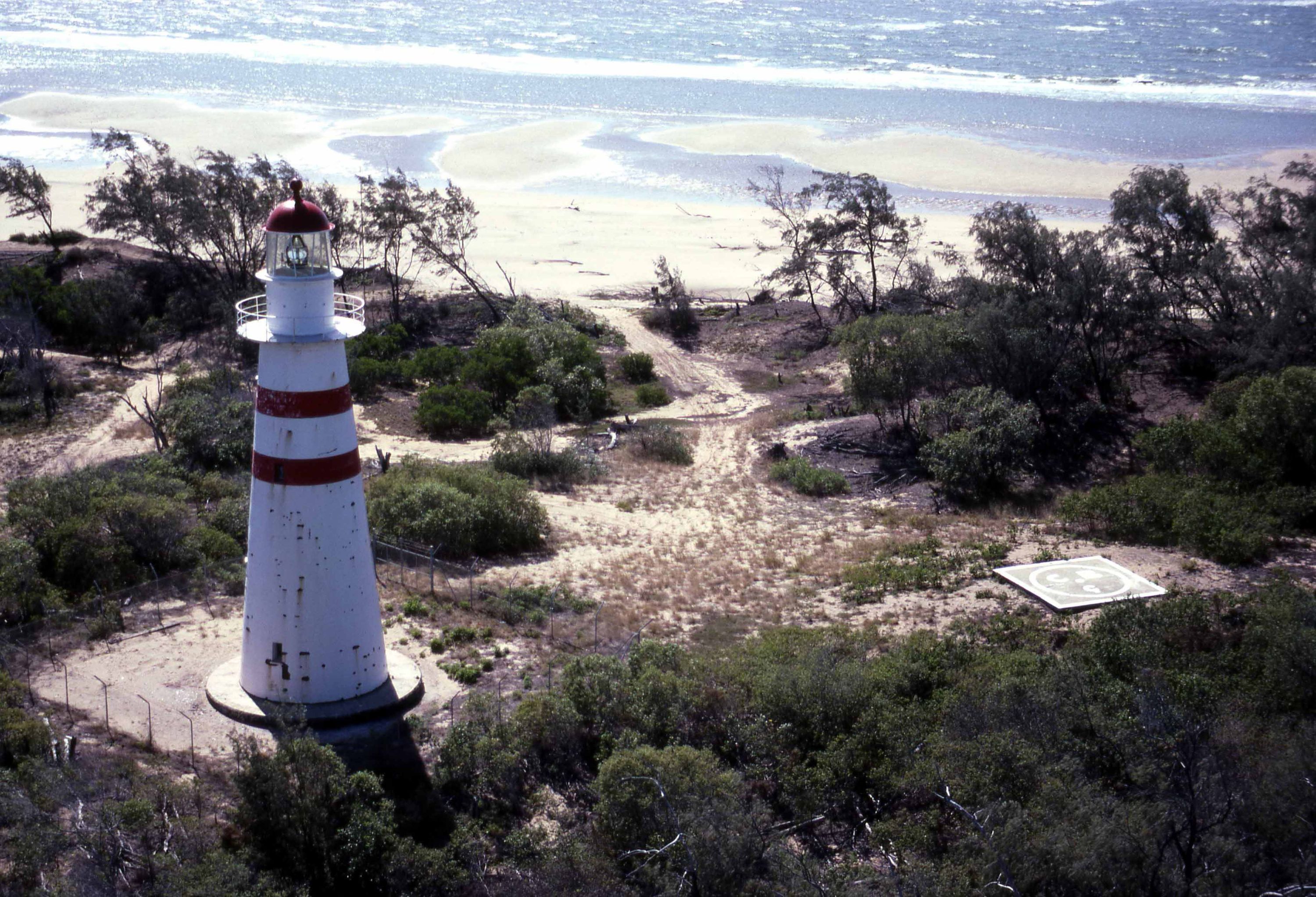 Photo showing alighthouse in profile surrounded by coastal scrub with a beach behind it.