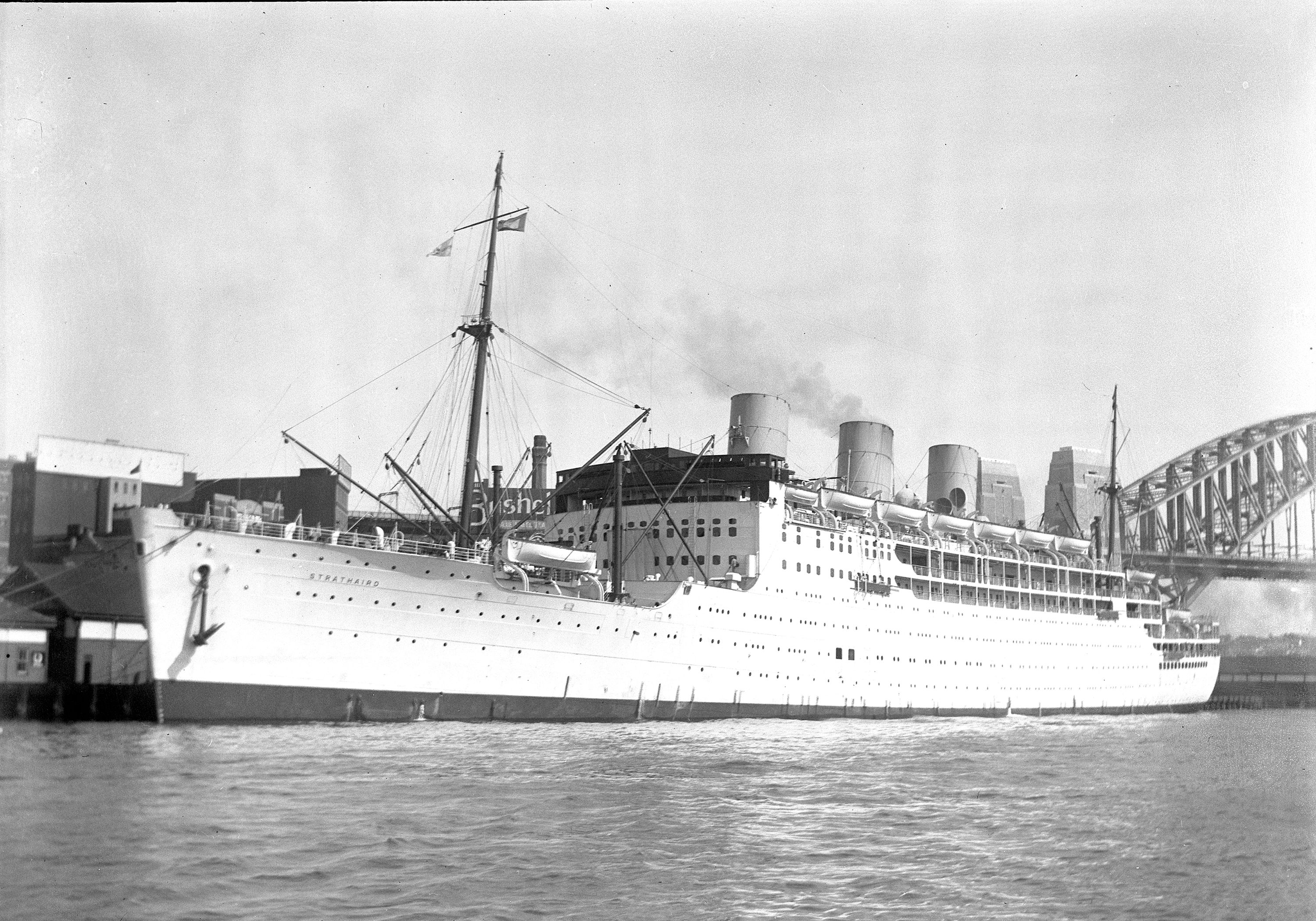 Black and white photograph of a large white cruise ship eith smoke coming from the funnels, at the dock in Sydney. The Harbour bridge is viable in the backgraound.