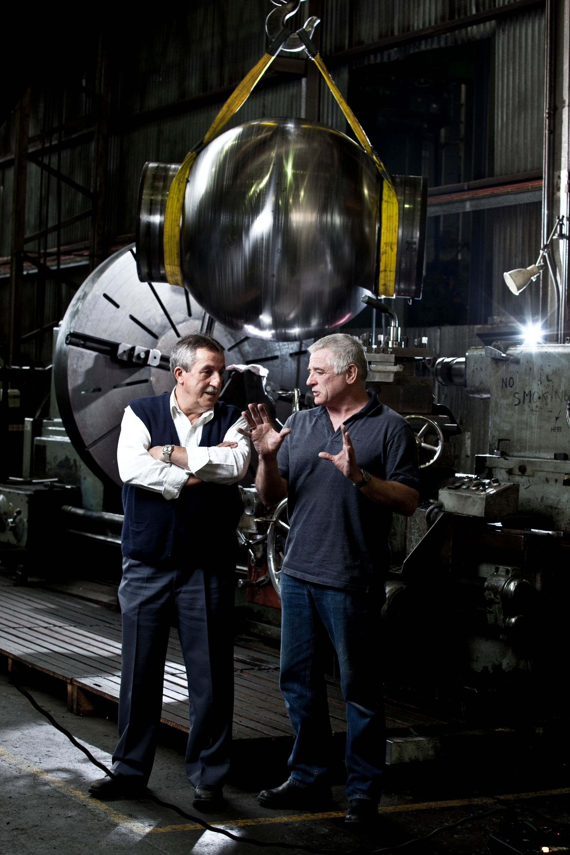 Two men talking inside a factory space behind them is a large, polished metal sphere susspended by yellow straps.