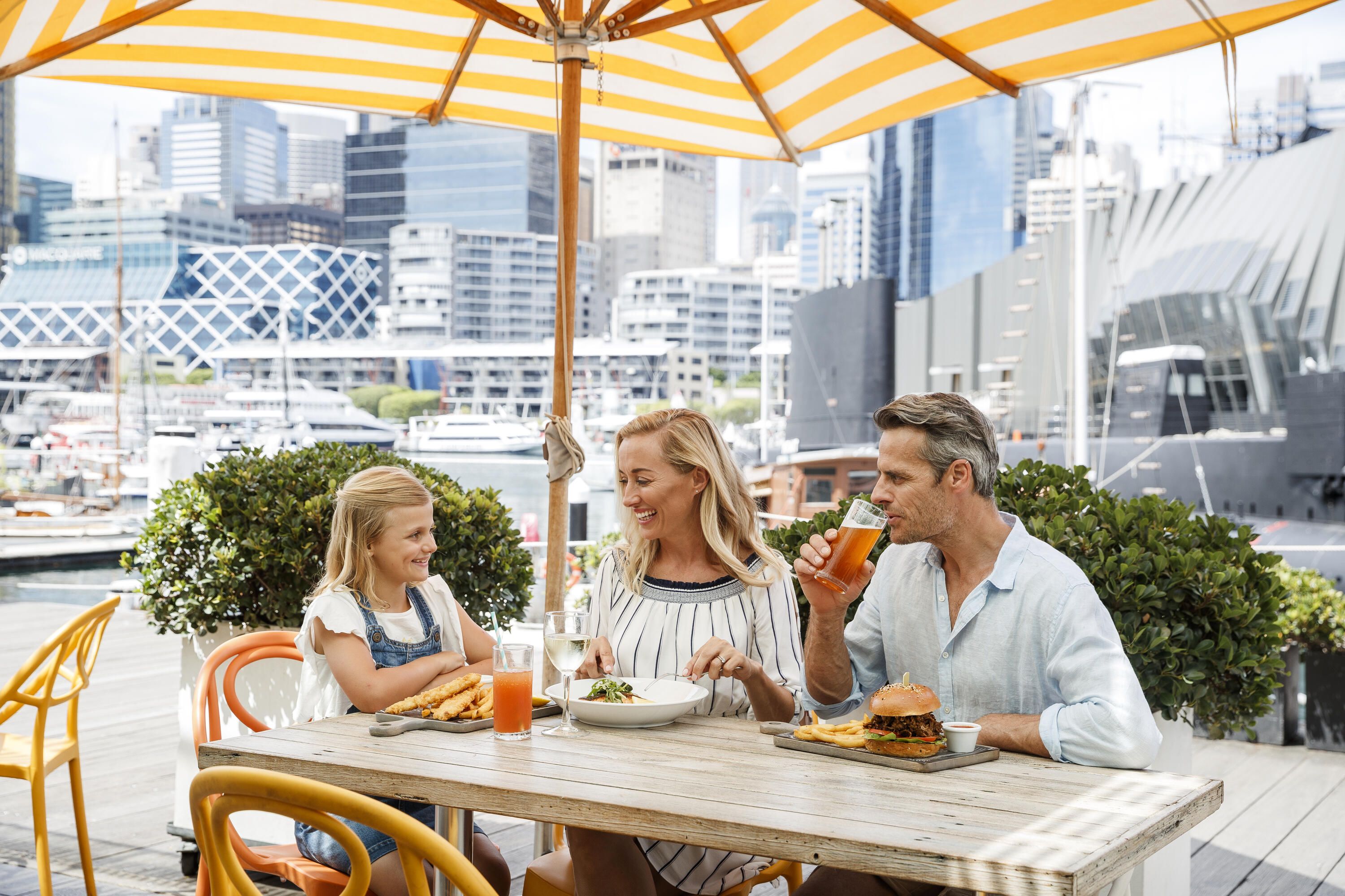 A Family with a mum dad and young girl are seated outdoor at the waterfront location enjoying a meal.
