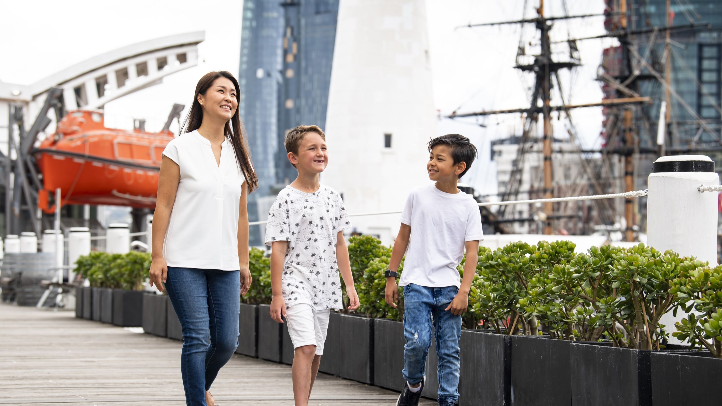 Photo of a women and two boys walking down a boardwalk, with shrubs, a lighthouse, tall ship masts and orange lifeboat in the background.
